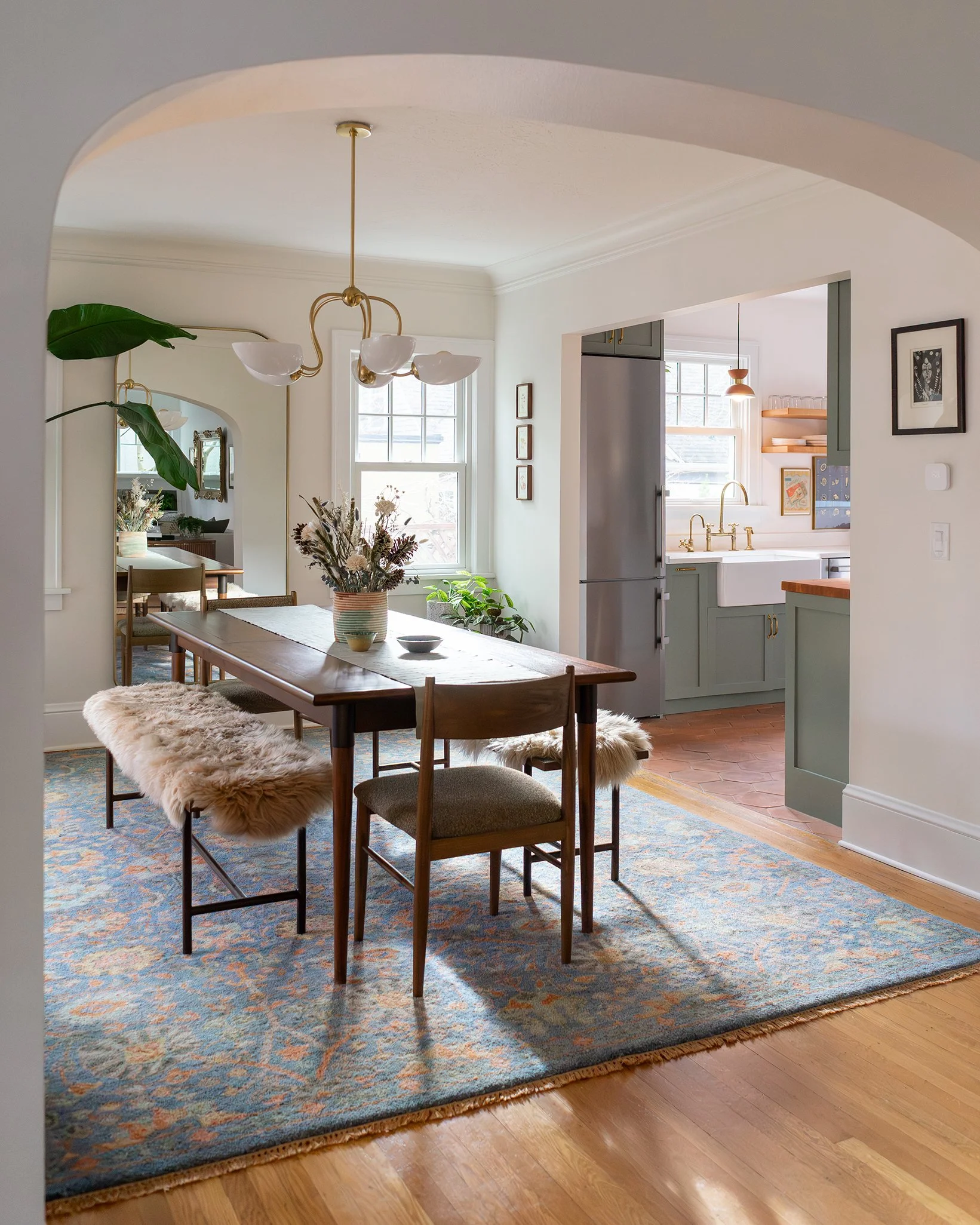 A dining area with a wooden table, four chairs, a fluffy sheepskin rug on one chair, and a large patterned area rug underneath. There are potted plants on the windowsill, a floral centerpiece on the table, and a ceiling light fixture with three white glass bowls. An open kitchen with green cabinets and gold hardware is visible in the background.