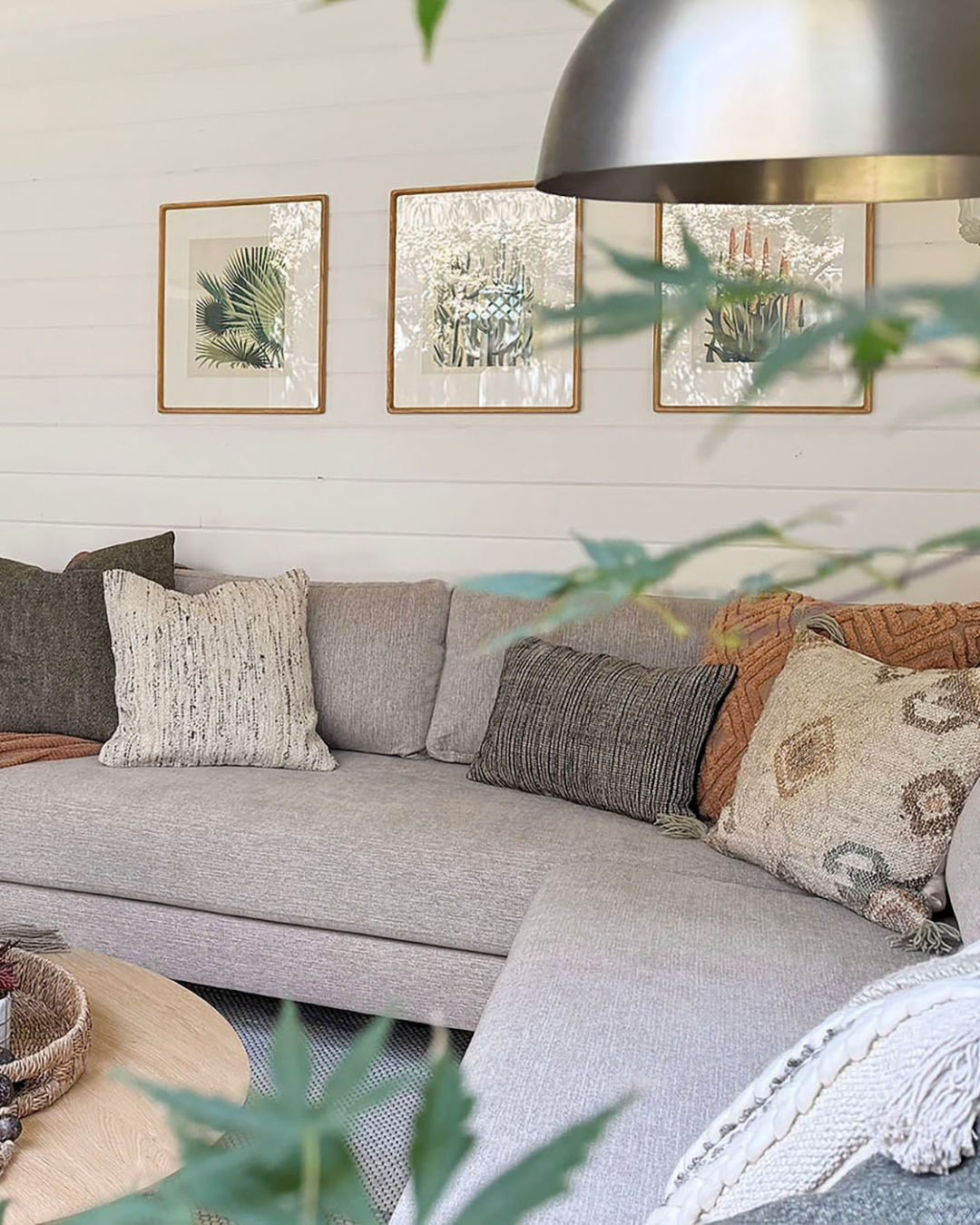 Living room with a light-colored L-shaped sofa decorated with various textured pillows. A wooden coffee table with a basket is in front of the sofa. The wall behind has three framed botanical prints, and a large metallic hanging lamp is partially visible. Green foliage is in the foreground.