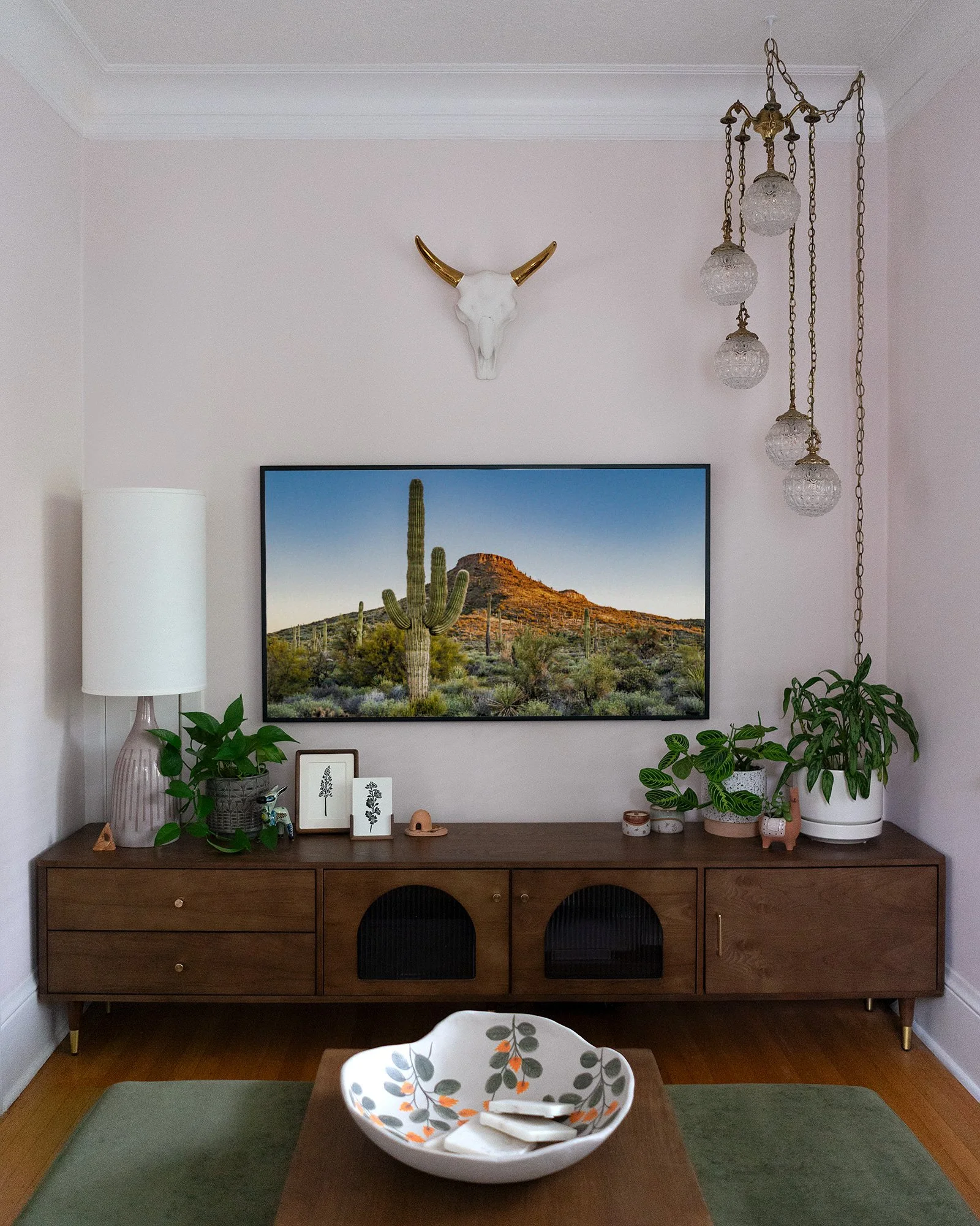 Living room with a wooden sideboard, potted plants, framed botanical art, wall-mounted cow skull with gold horns, large framed desert landscape photo, white table lamp, hanging glass globe chandelier, and a table with a decorative bowl and utensils.