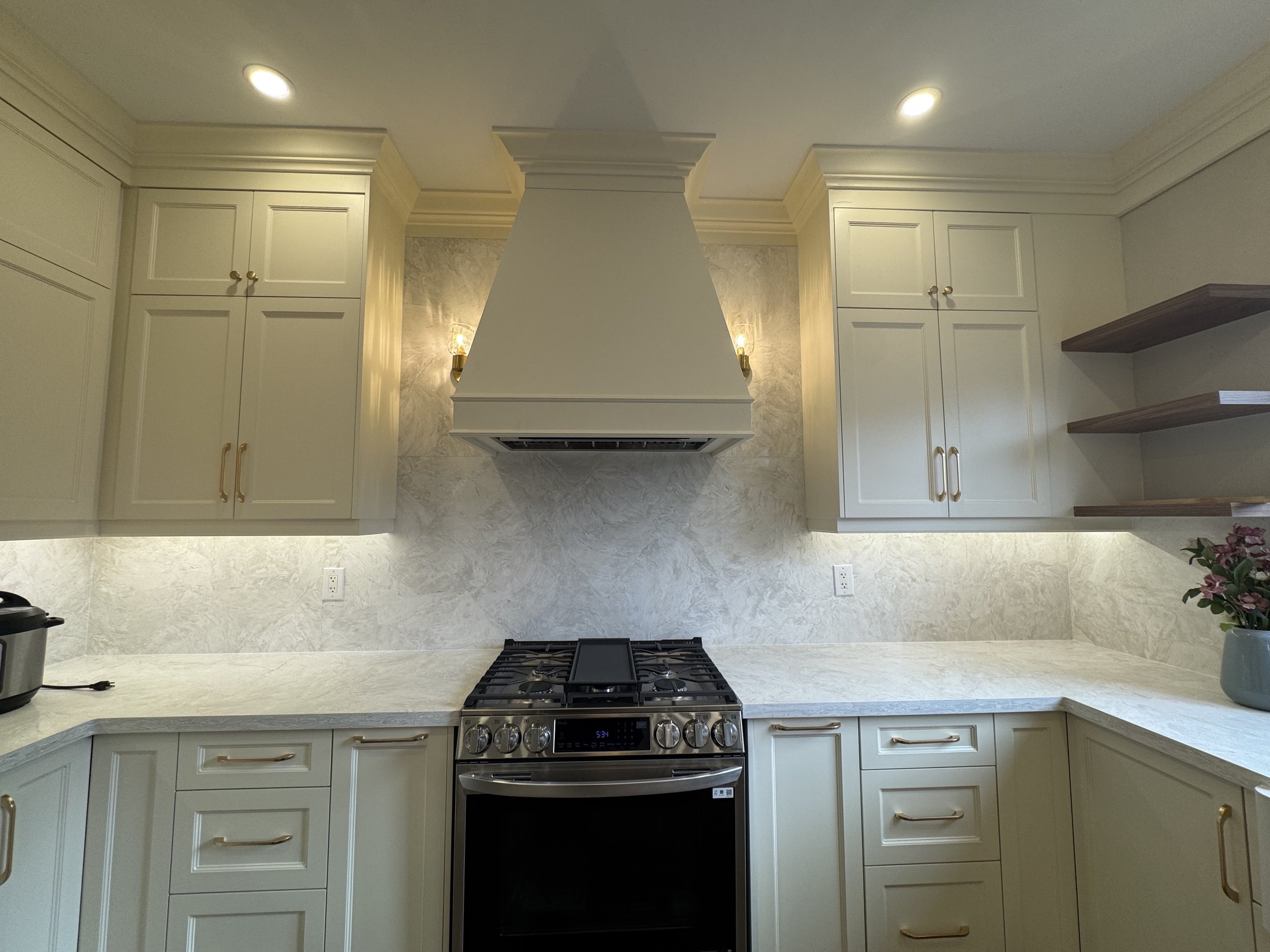 A kitchen with cream-colored cabinetry, granite countertops, a white range hood, a stove, and open wooden shelves on the right. There is a potted plant with pink flowers on the countertop to the right.