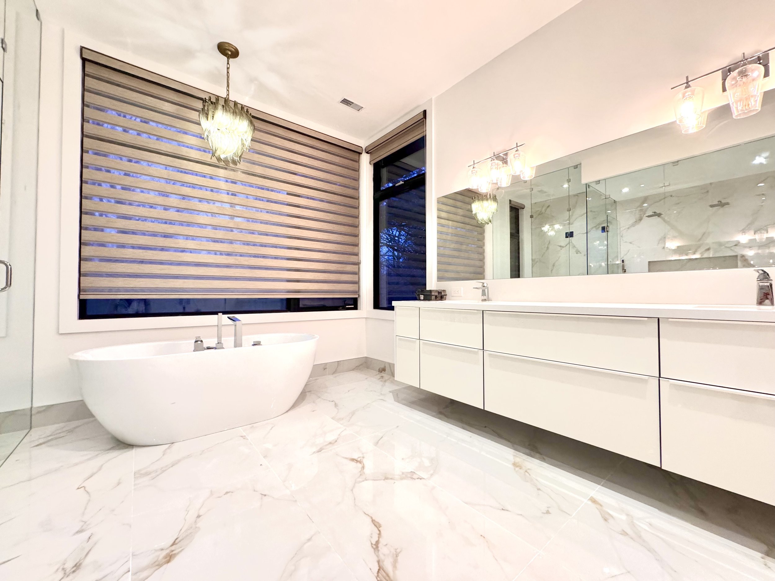 Modern bathroom with a white freestanding bathtub, marble floor, large mirror, and white cabinetry, illuminated by decorative light fixtures.