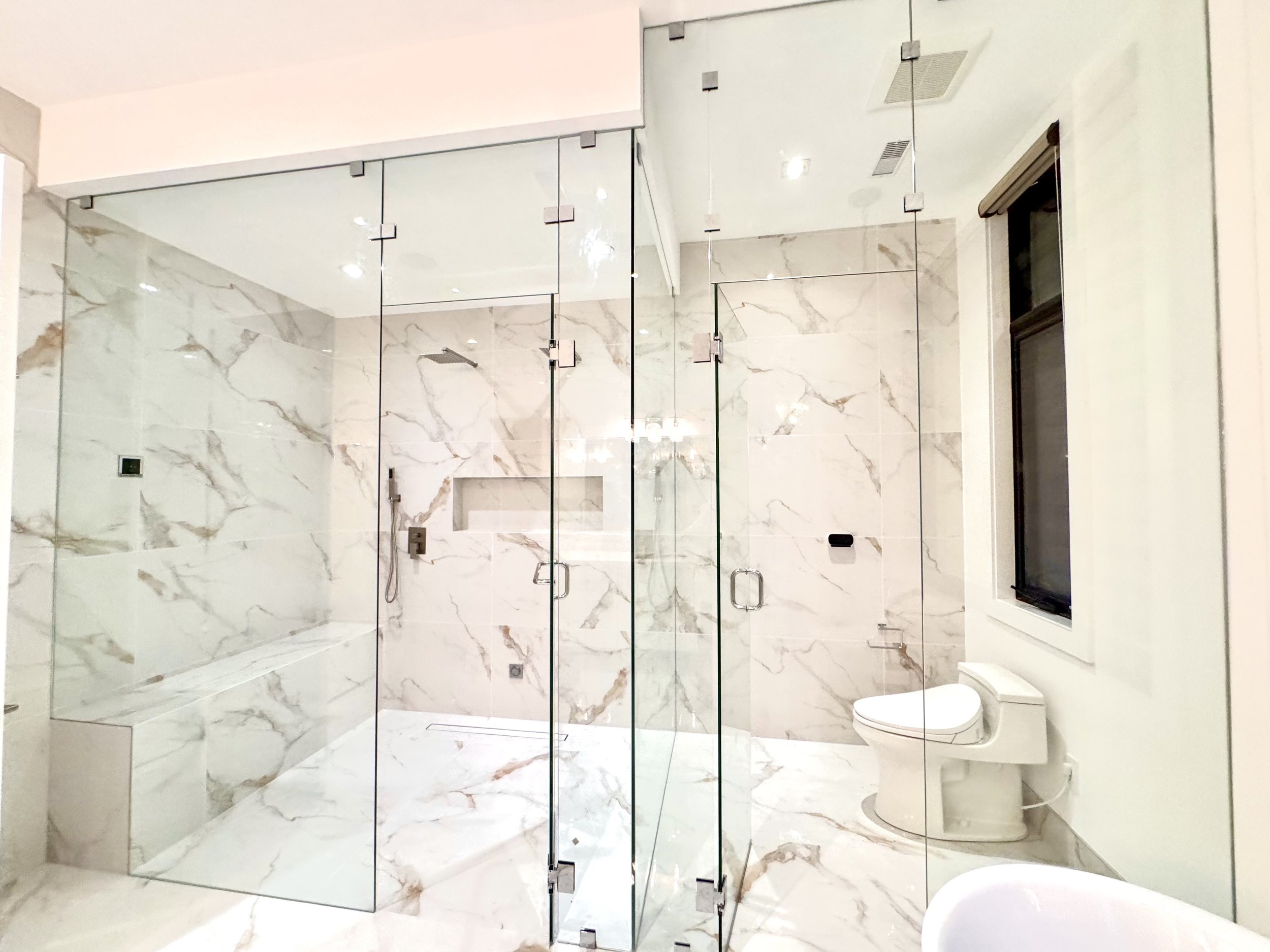 Modern bathroom with glass shower enclosure, marble tile walls and floor, a built-in bench in the shower, a window, and a wall-mounted toilet.