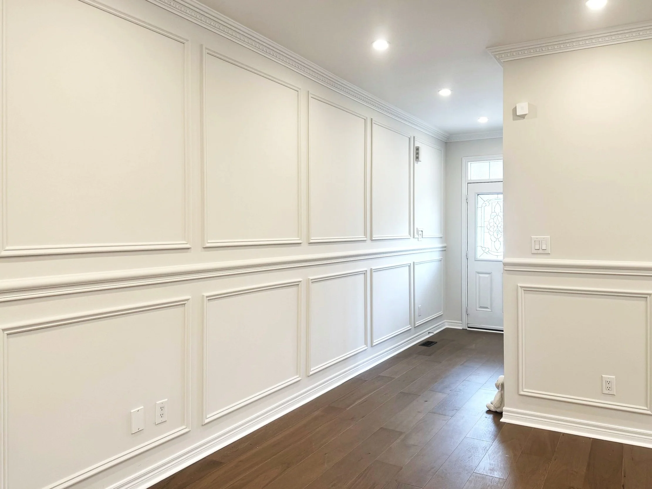Interior view of a hallway with white wall paneling, crown molding, hardwood flooring, and a front door with decorative glass.