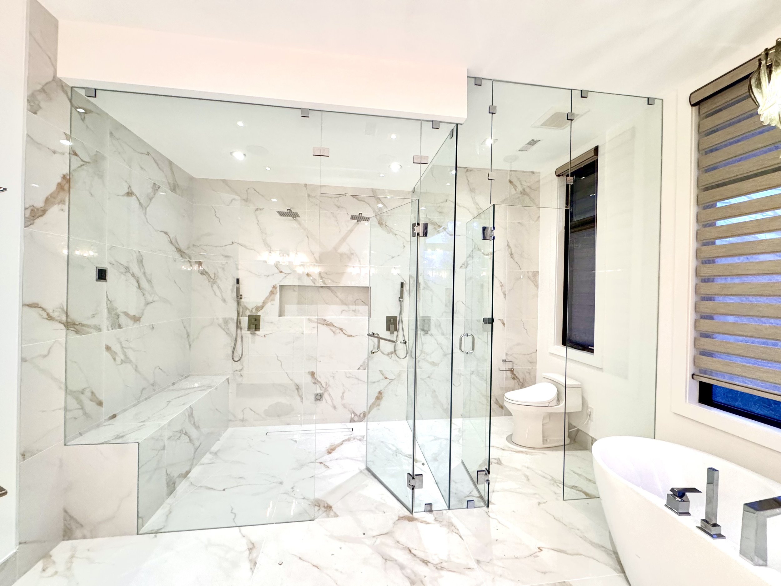 Modern bathroom with marble walls and floors, glass shower enclosure, freestanding bathtub, toilet, and window with blinds.