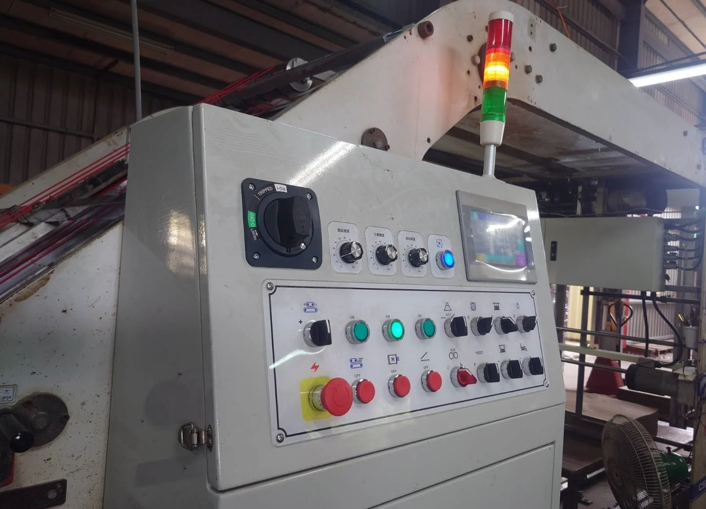 Industrial control panel with switches, buttons, a display screen, and a traffic light signal above it, installed in a factory setting.