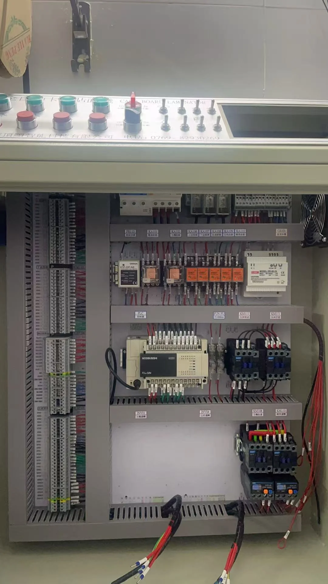 Electrical control panel with switches, relays, and wiring, mounted on a wall with a grey enclosure.