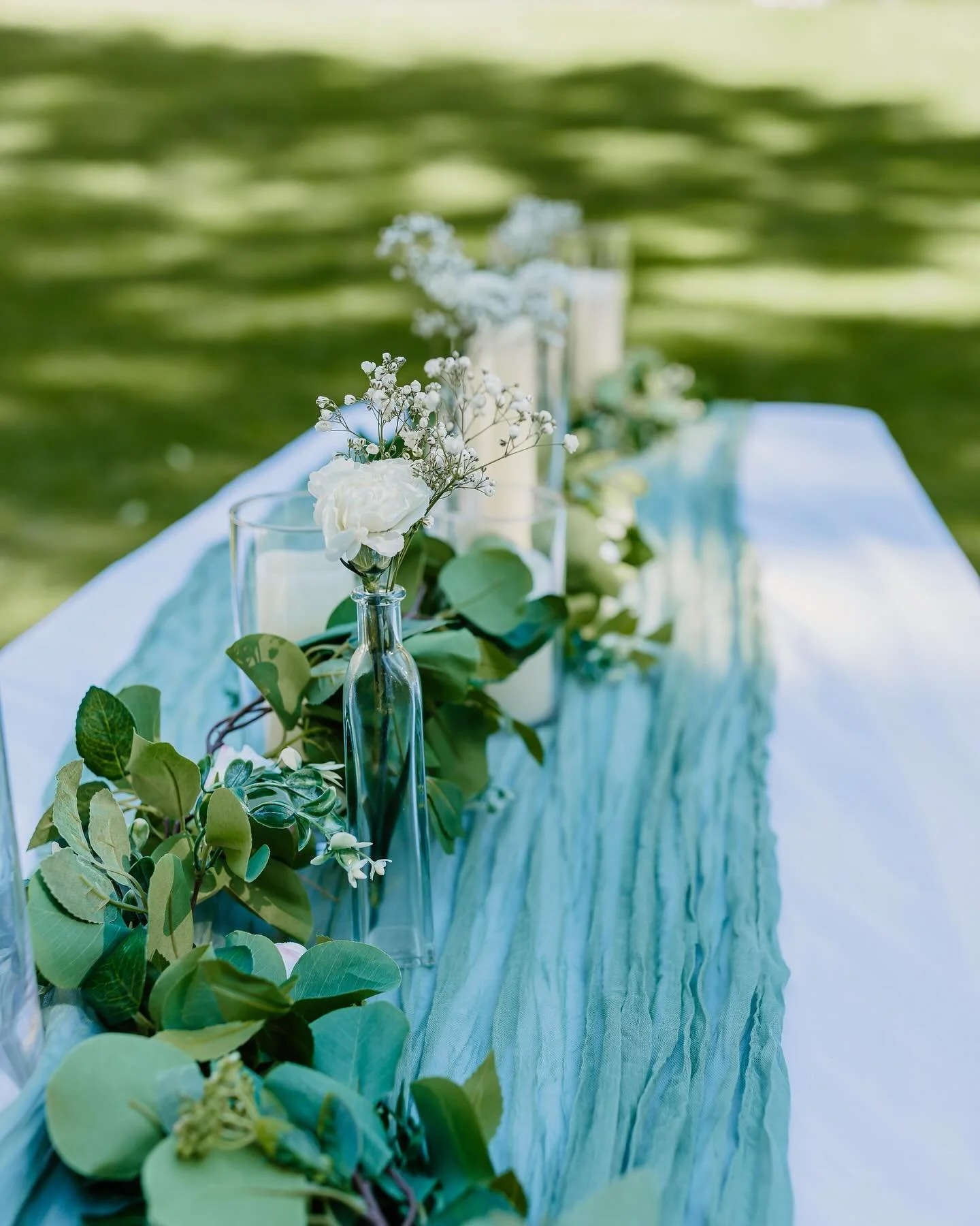 Decorative table setup with white flowers, green leaves, tall glass candle holders, and a light blue fabric runner outdoors.