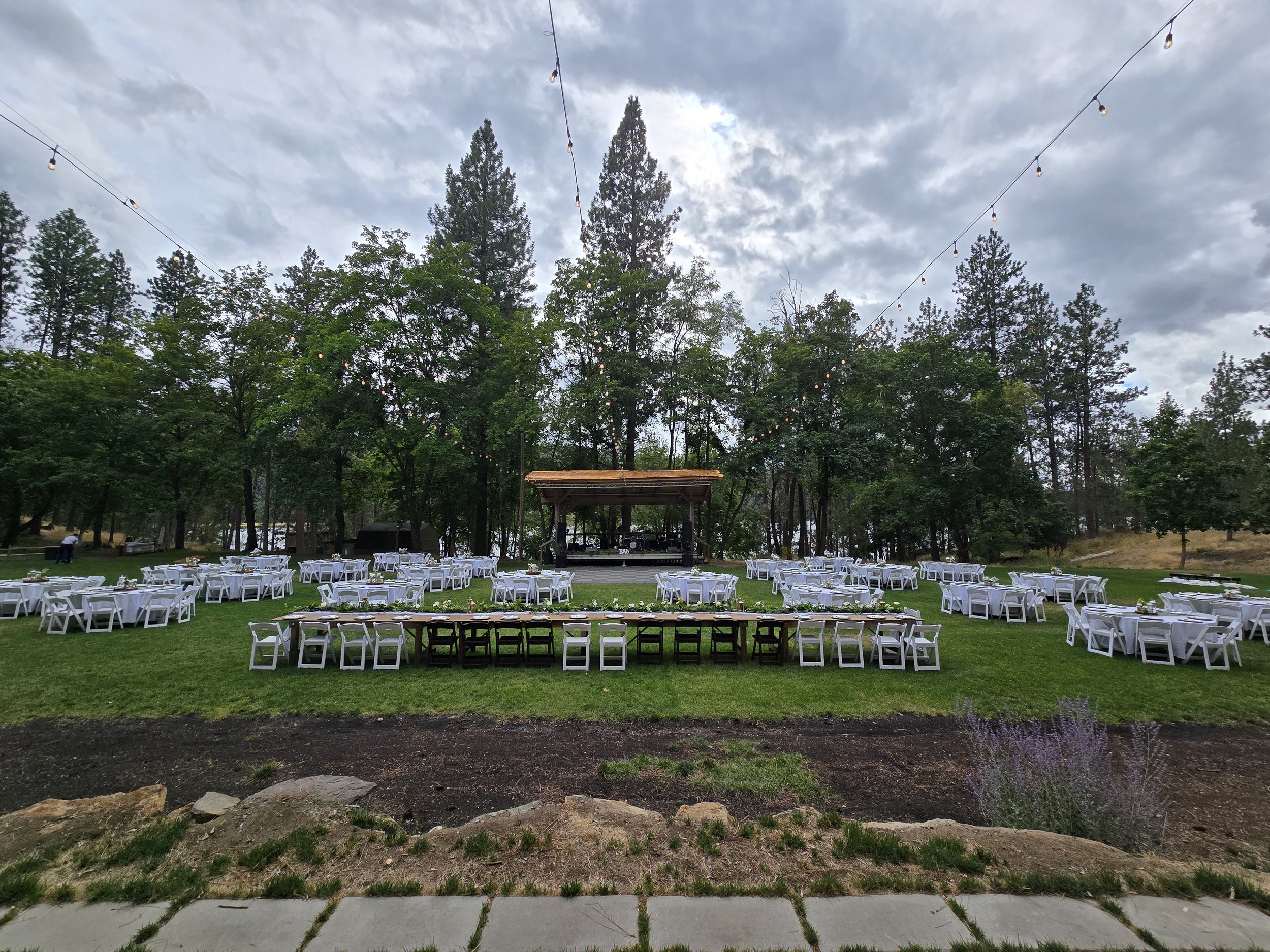 Outdoor event setup with white tables and chairs on a grassy lawn, a stage with a wooden roof in the background, string lights hanging above, and trees surrounding the area under a cloudy sky.