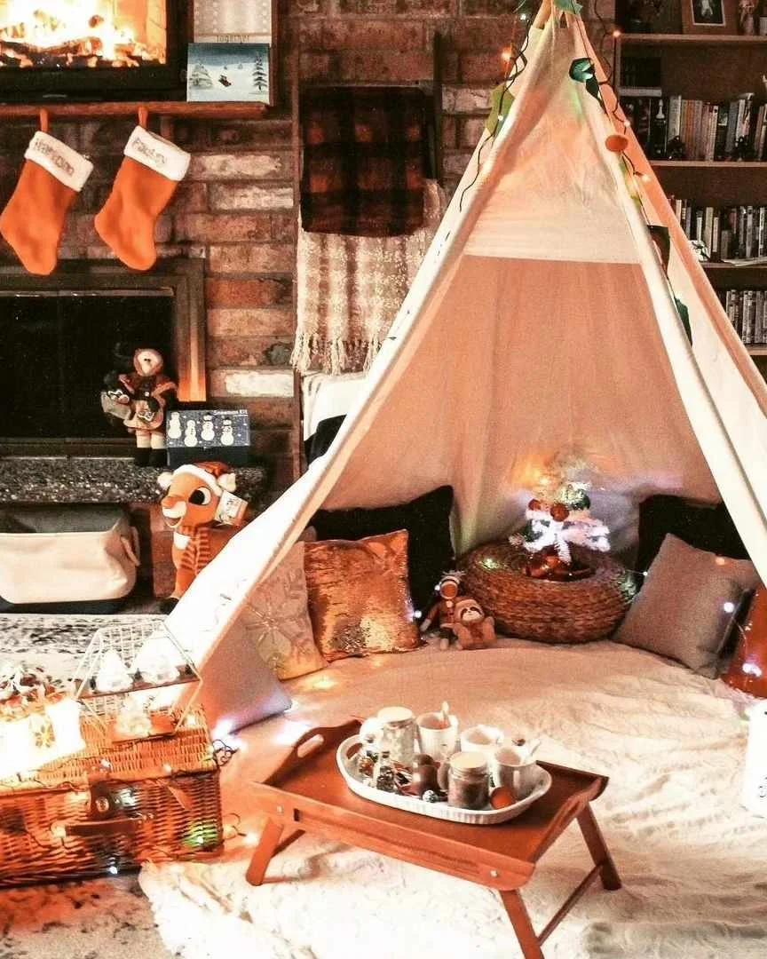 A cozy living room with Christmas decorations, including two hanging stockings over a fireplace, a small teepee tent with plush pillows and string lights, and a table with mugs and holiday treats. There are also stuffed animals and Christmas ornament