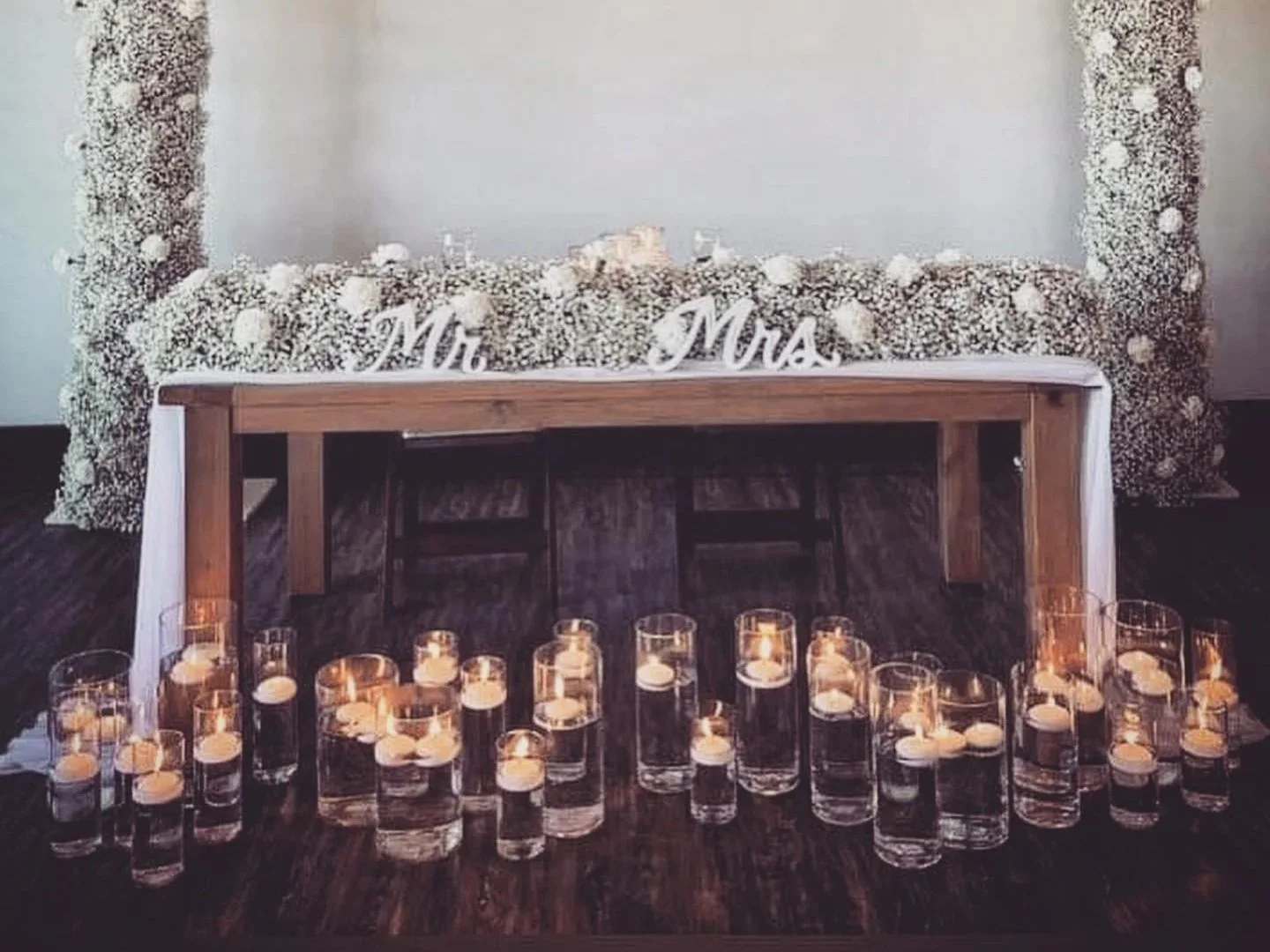 A wedding setup with a floral backdrop, 'Mr. & Mrs.' signs on a table, candles in glass holders on the floor.