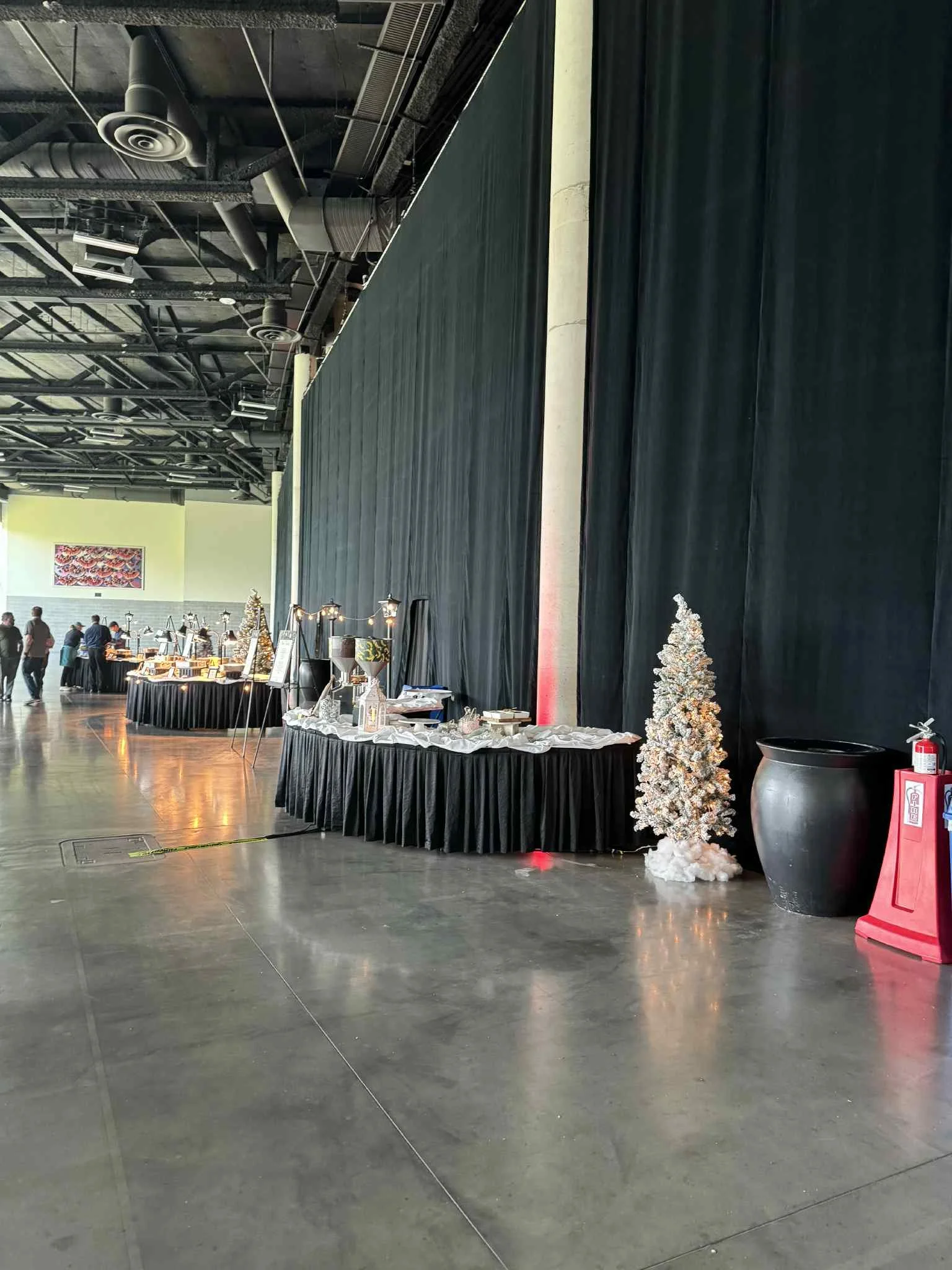 Indoor event space with black curtains and tables decorated for a holiday or Christmas celebration, featuring a small white Christmas tree, large black pot, fire extinguisher, and people in the background.