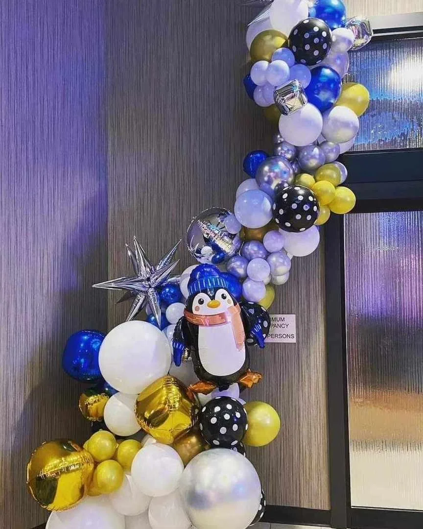 Colorful balloon arch with black, white, gold, and blue balloons, including polka-dot and metallic designs, featuring a penguin-shaped balloon with a pink scarf.