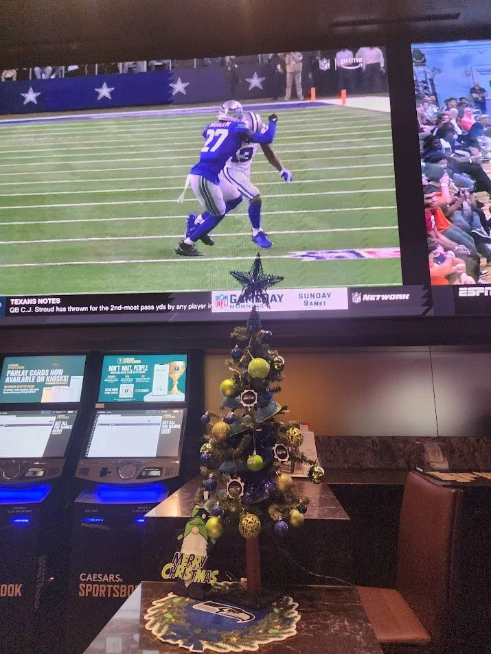 A large television screen displaying a football game, with a player in a blue uniform catching the ball while being tackled. Below the screen, there are lottery machines, a small decorated Christmas tree with blue, green, and gold ornaments, and a Se