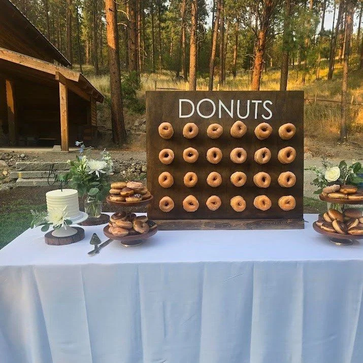 A table with a white tablecloth displays donuts on a wooden board labeled "DONUTS," with some donuts on plates and decorative flowers placed around.