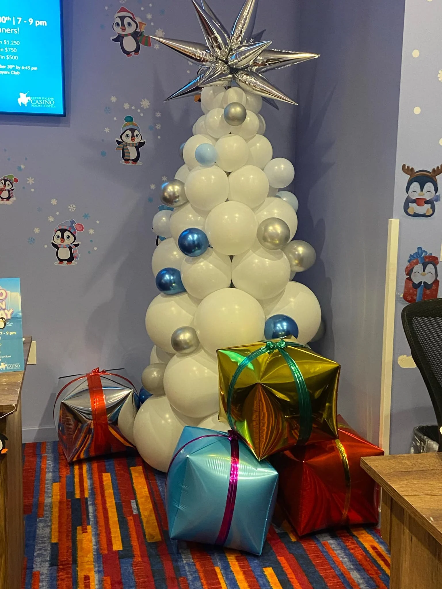 Decorative Christmas tree made of white and silver balloons with a large silver star on top, surrounded by colorful wrapped presents, in a festive room with holiday themed wall stickers featuring penguins and reindeer.