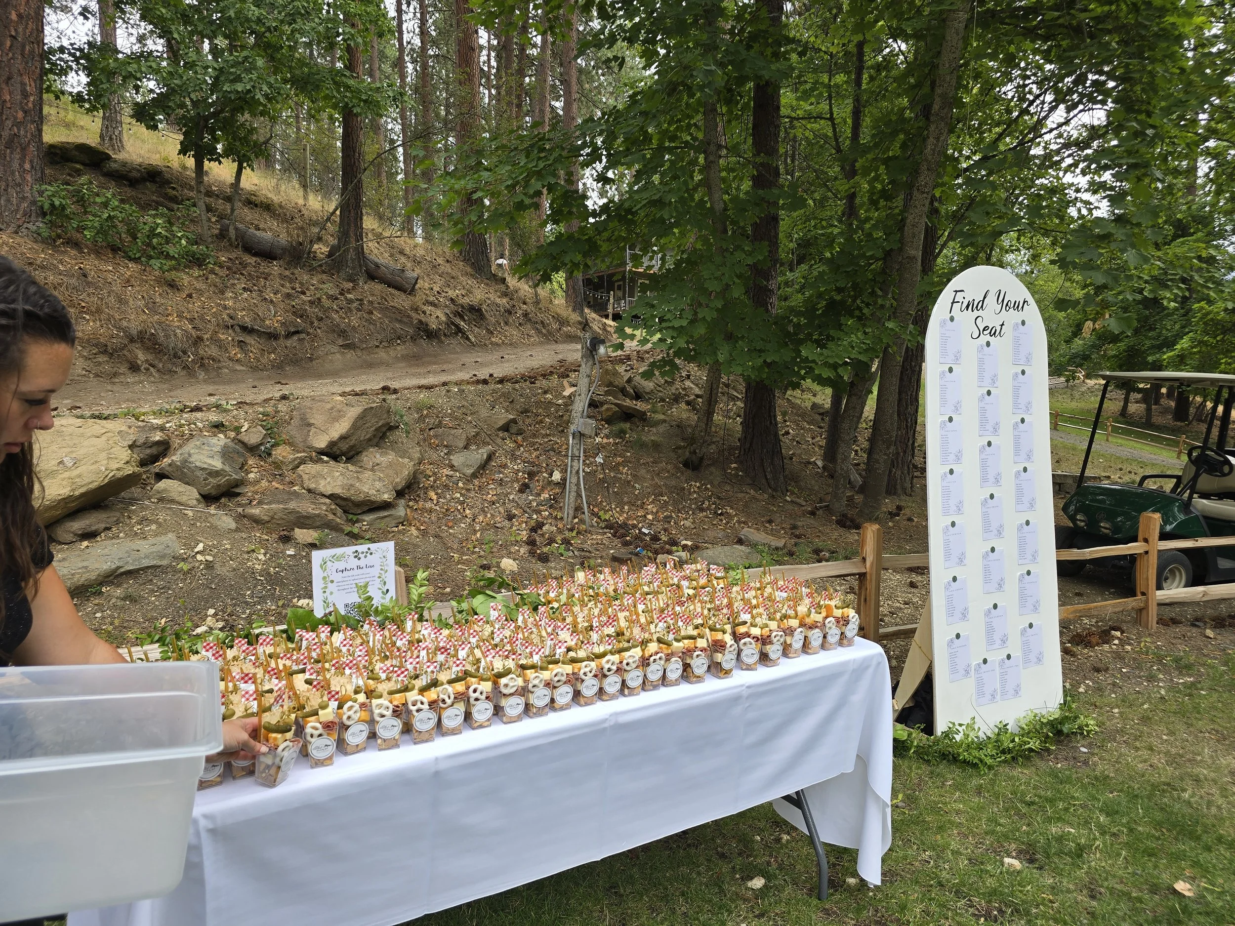 A table with small jars and decorations set up outdoors in a wooded area, with a large seating chart on a white board nearby that says 'Find Your Seat' at a gathering or event.