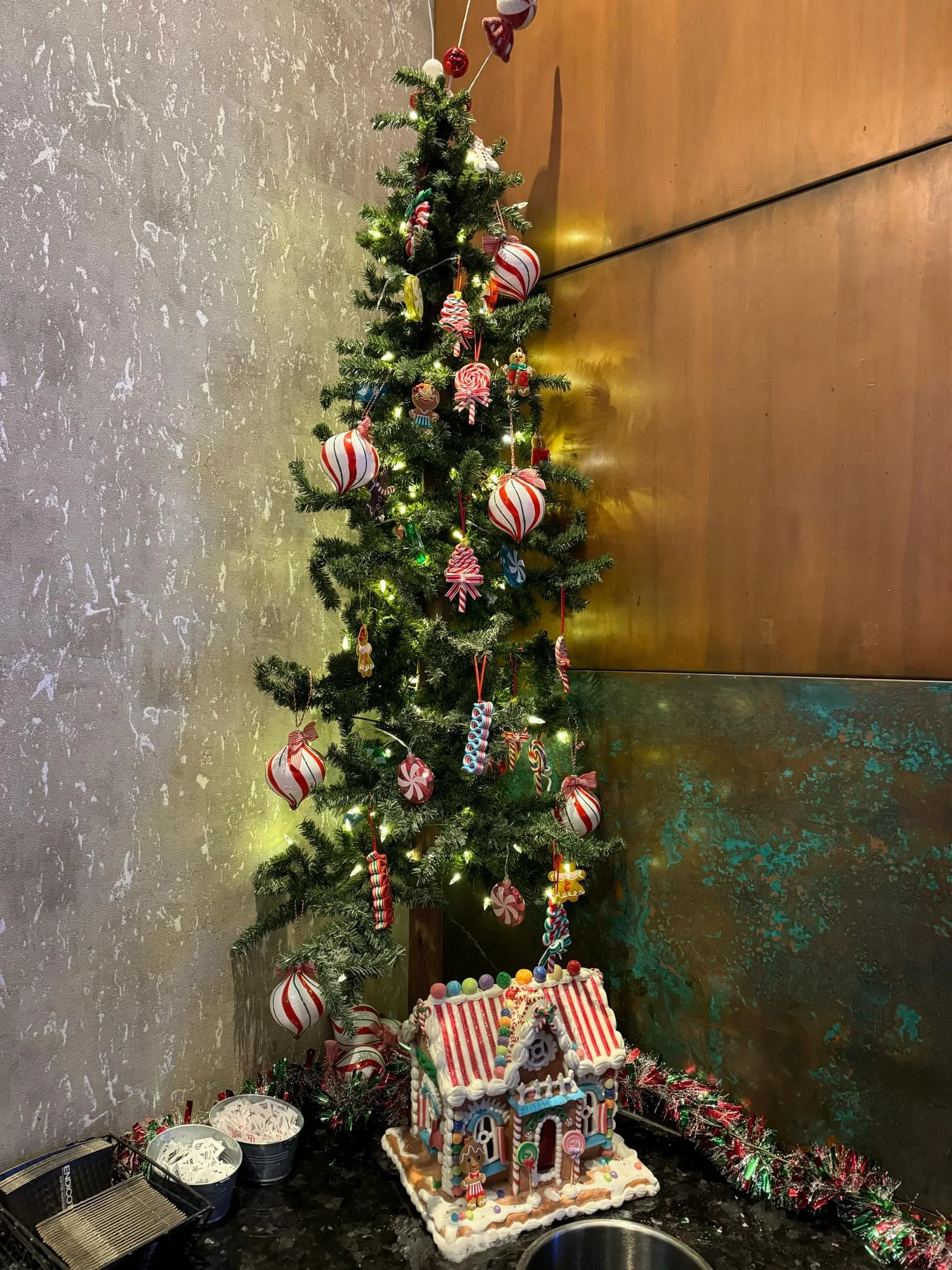 A decorated Christmas tree with red, white, and green ornaments, candy ornaments, and lights. The tree stands next to a gingerbread house decorated with candies, surrounded by tinsel and small bowls.