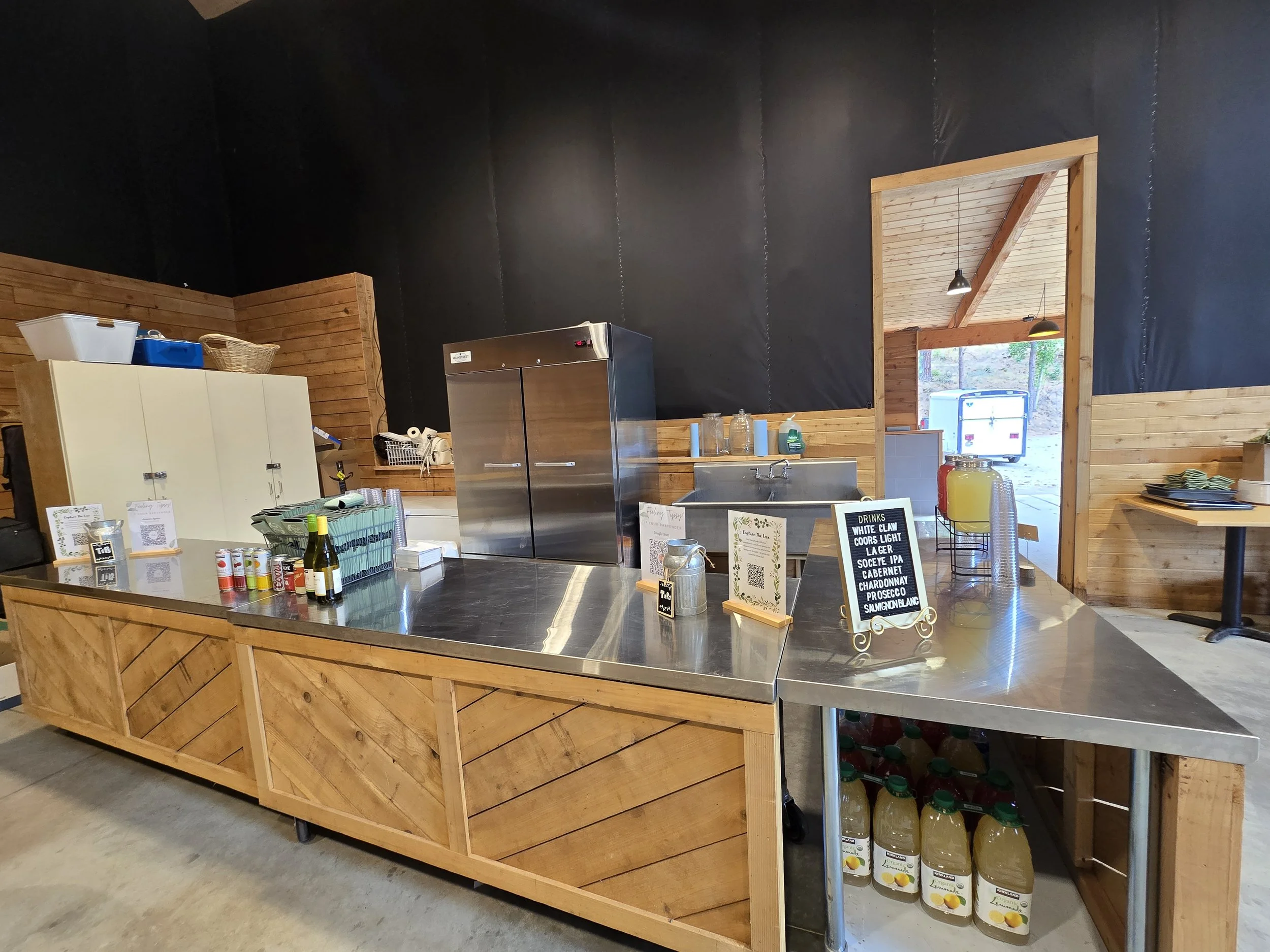 A rustic refreshment area with a wooden counter, a stainless steel refrigerator, and bottles of juice on a shelf below. There are three informational signs and a menu board with drinks listed, including beers and wine. The space has wood-paneled wall