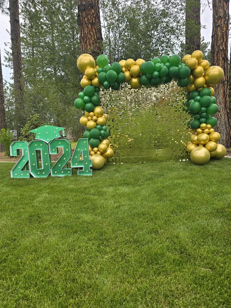 Green and gold balloon arch with a mirrored backdrop, and illuminated "2024" sign with a graduation cap, on a grassy area surrounded by trees.
