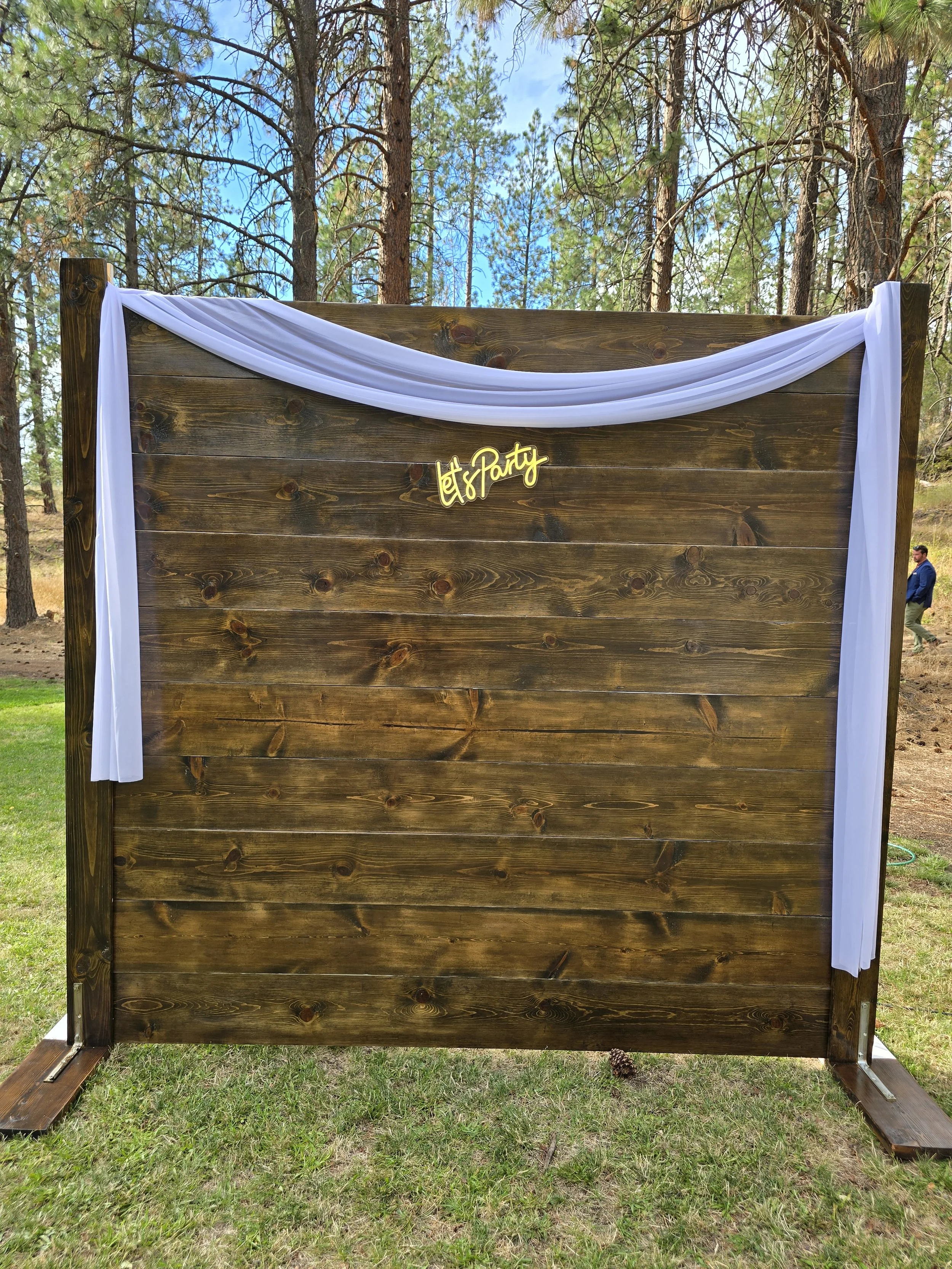 A wooden photo backdrop with white drapes on both sides and a yellow neon sign that reads "Let's Party" in the center, set outdoors with trees and grass in the background.