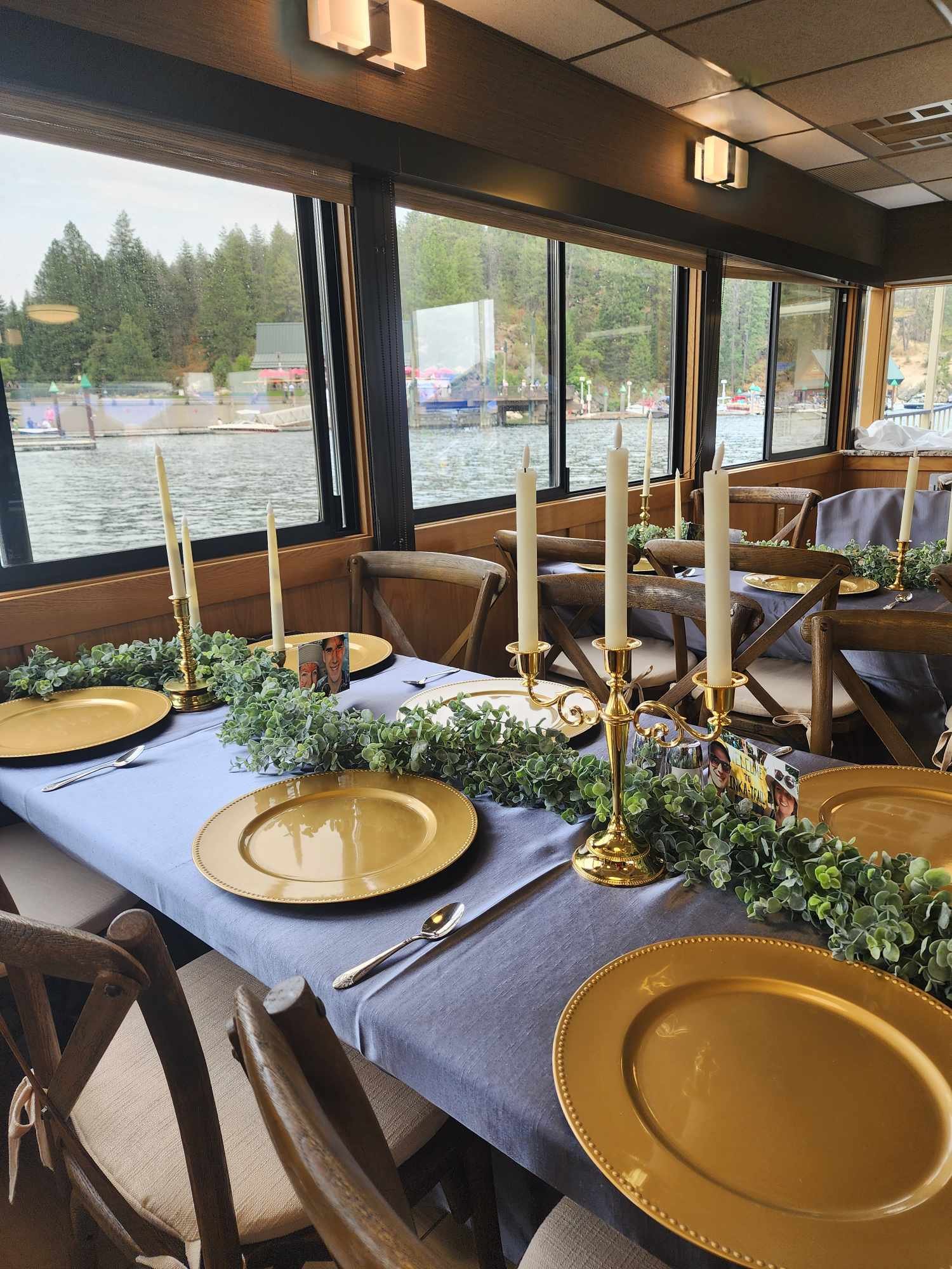 A beautifully decorated dining table set with gold plates, silverware, and tall white candles in gold candle holders. The table is adorned with green foliage, and there are small photographs on the table. The room has large windows overlooking a body