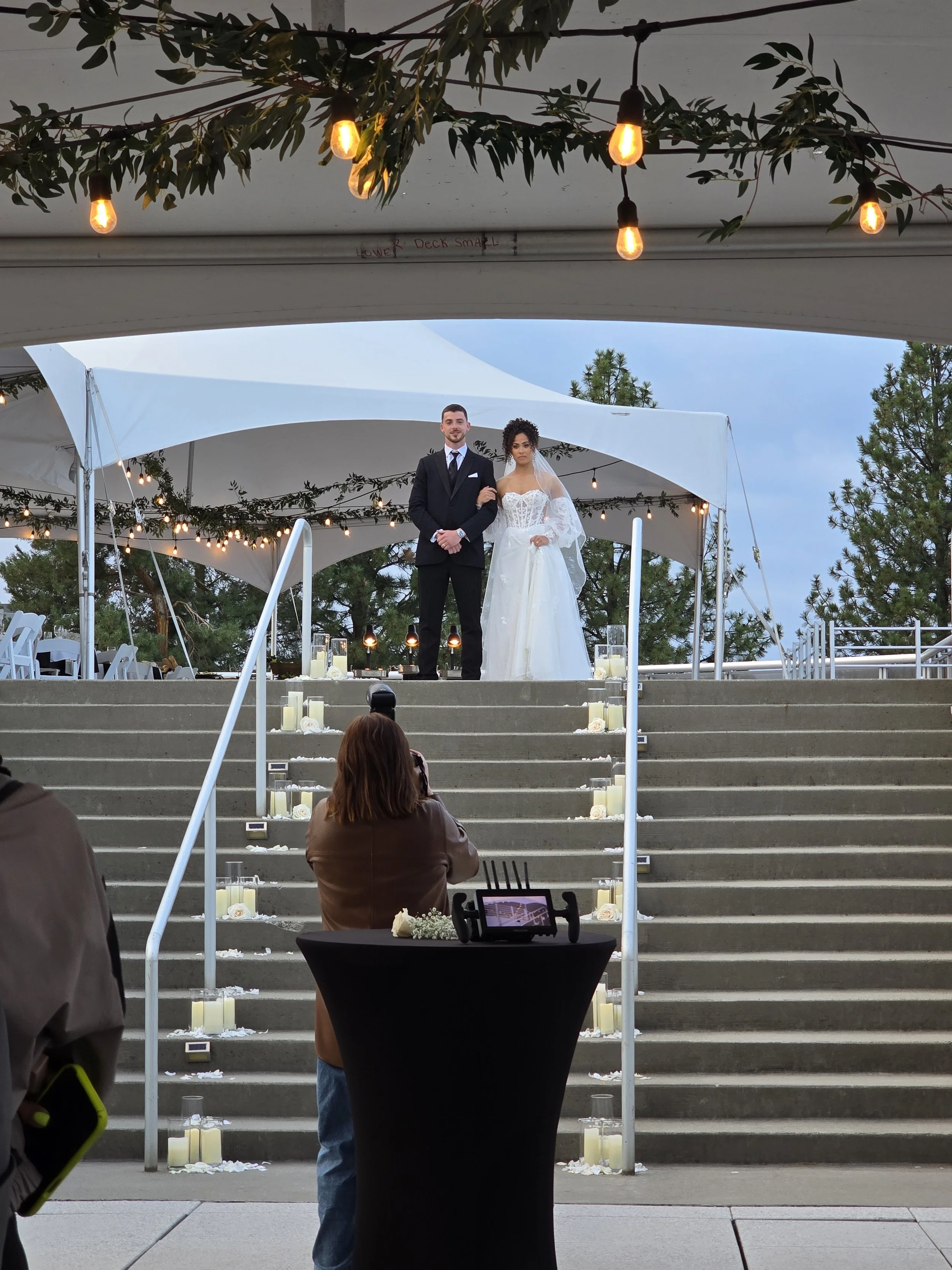 A bride and groom standing on a raised platform under a white canopy during an outdoor wedding, with guests seated below and candles lining the steps, with trees and a cloudy sky in the background.