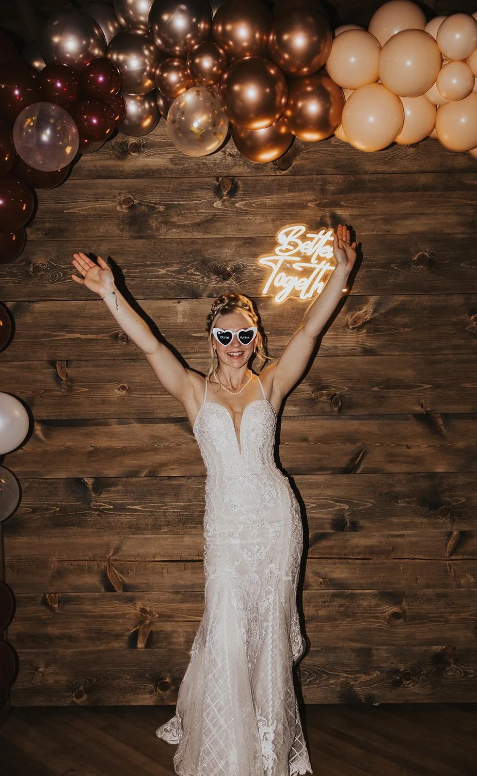 A woman in a wedding dress with sunglasses posing with arms raised, standing in front of a wooden wall decorated with balloons and a neon sign that says "Better Together."