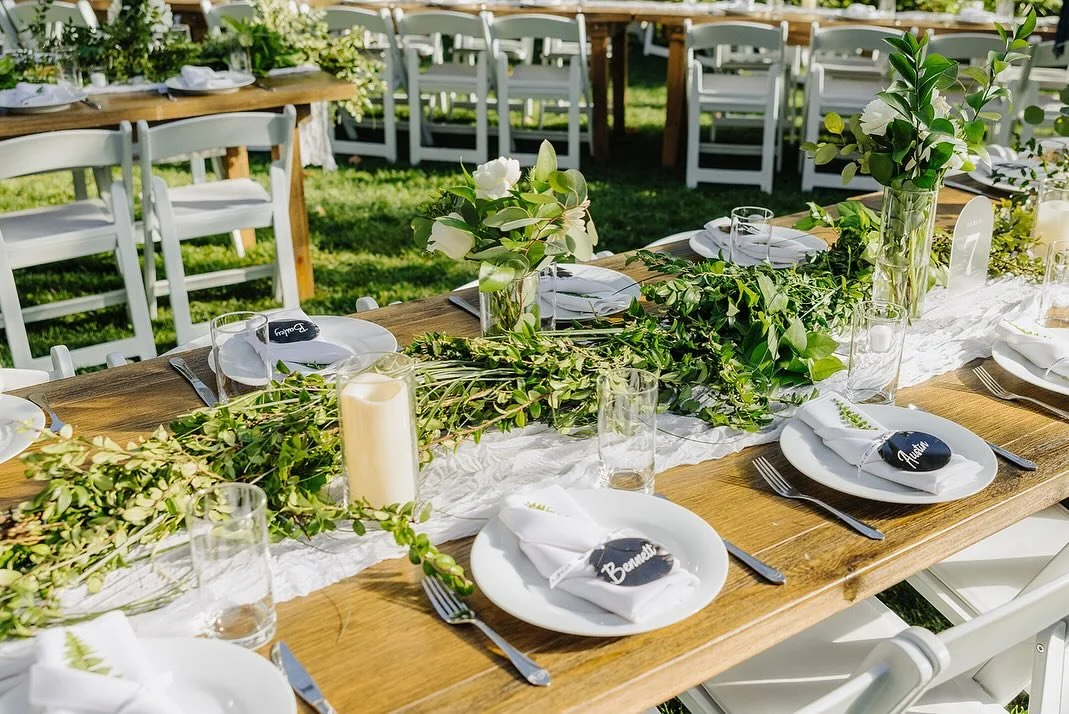 Outdoor table set for a celebration with white dishes, napkins labeled "Beauty" and "Groom," glassware, and green foliage centerpieces under sunlight.