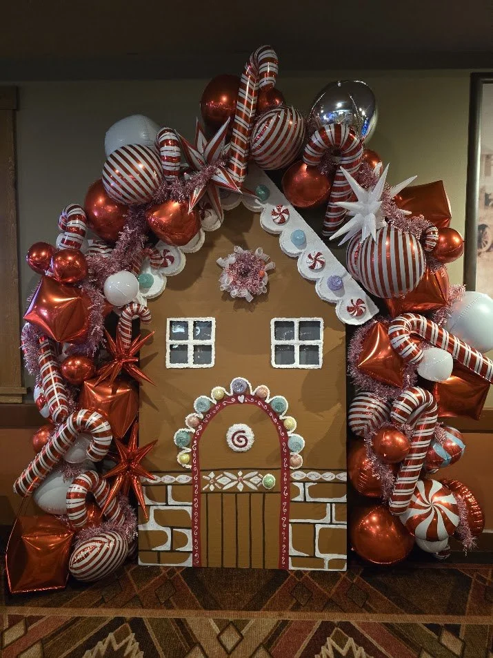 Decorative gingerbread house with red, white, and silver balloons, candy canes, stars, and peppermint candies for a festive holiday display.