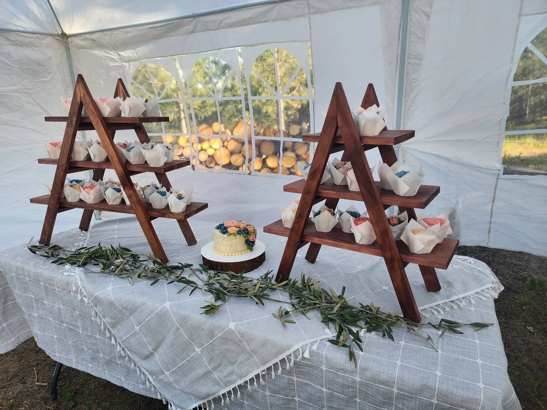 Display table with cupcakes on wooden stands, small decorated cake, and greenery in a white tent with wood logs outside.