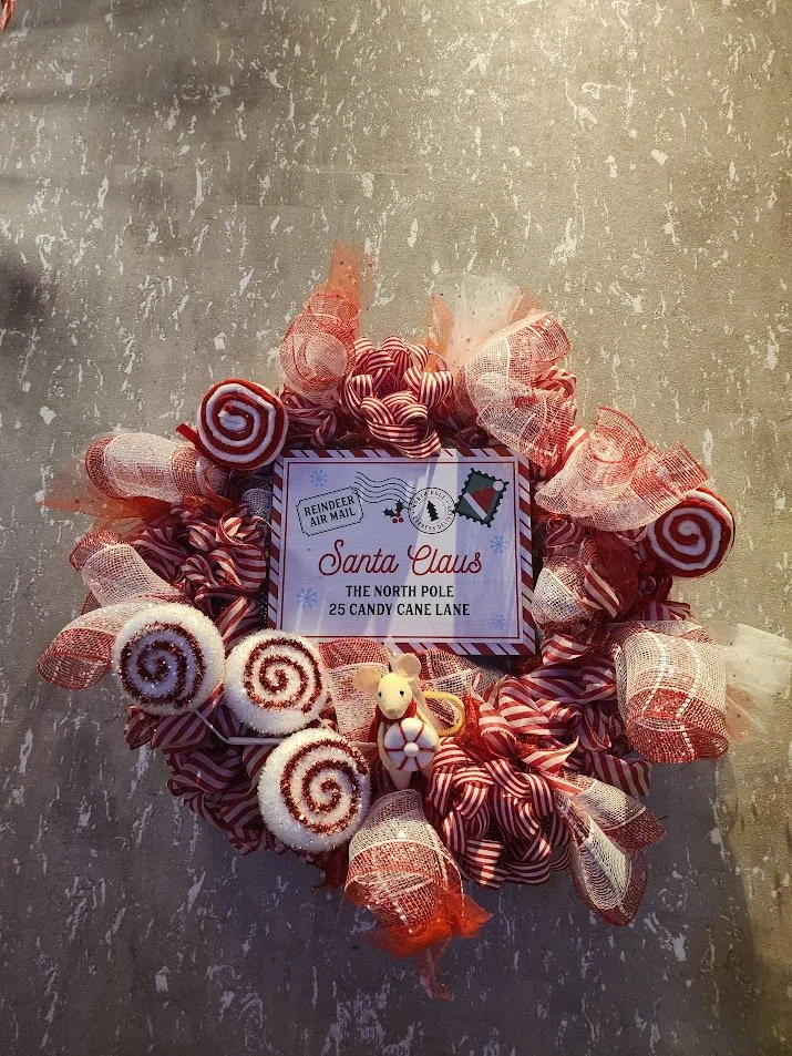 Christmas wreath decorated with red and white ribbon, candy cane-shaped lollipops, and a small reindeer figurine, with a note addressed to Santa Claus from the North Pole.