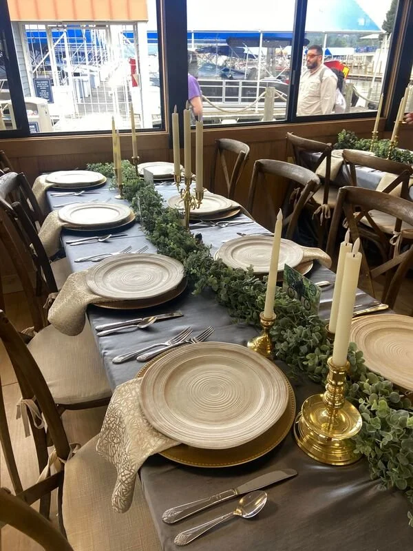 A table set for a formal event with a dark tablecloth, elegant gold candlesticks with tall white candles, transparent chargers, cream-colored napkins, and silverware, positioned in front of large windows overlooking a marina with boats and a walkway.