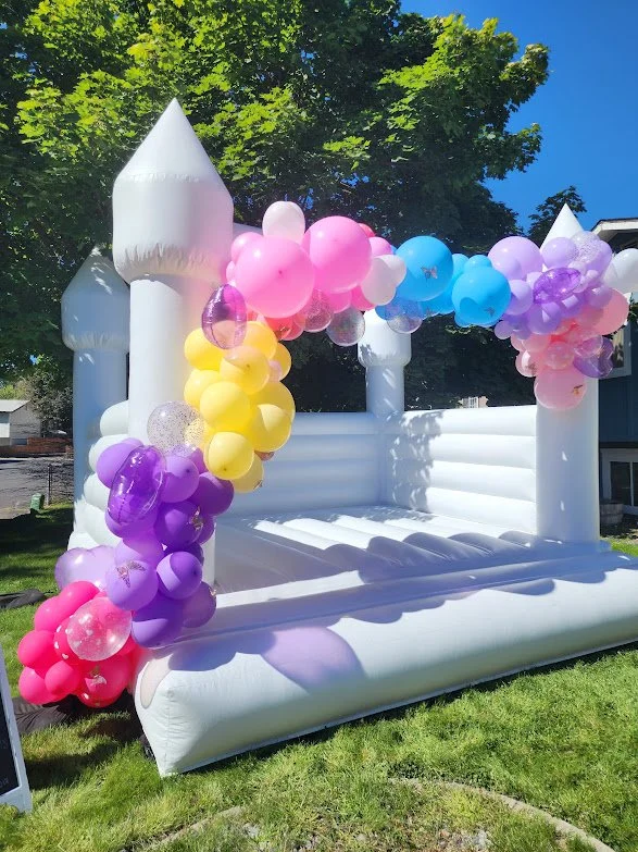 Inflatable castle decorated with colorful balloons including pink, purple, yellow, blue, and white, set outdoors on grass with trees and a blue sky in the background.