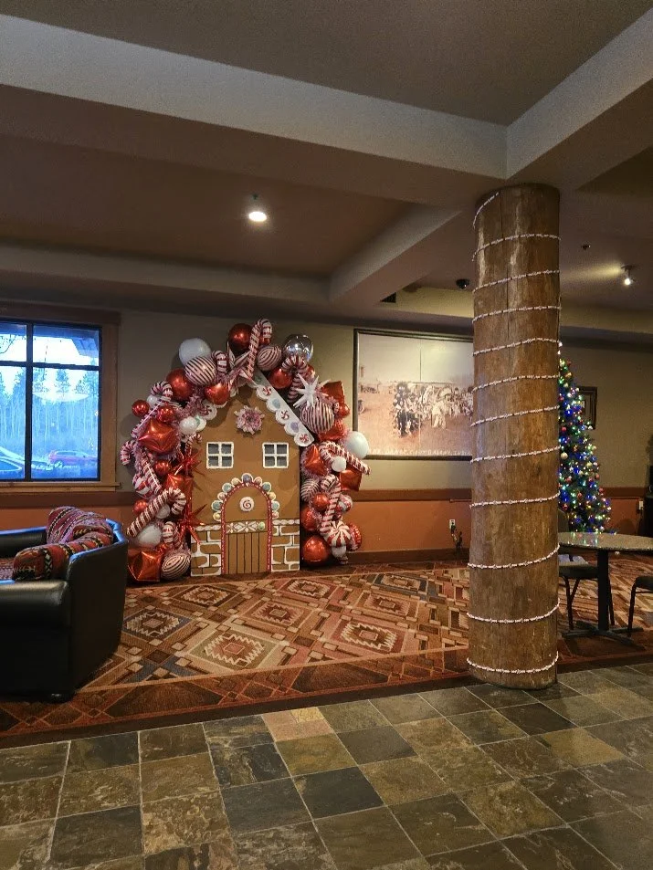 Holiday display featuring a gingerbread house backdrop decorated with red, white, and silver ornaments and candy canes, with a winter scene painting, a Christmas tree with lights, and a wrapped pillar.