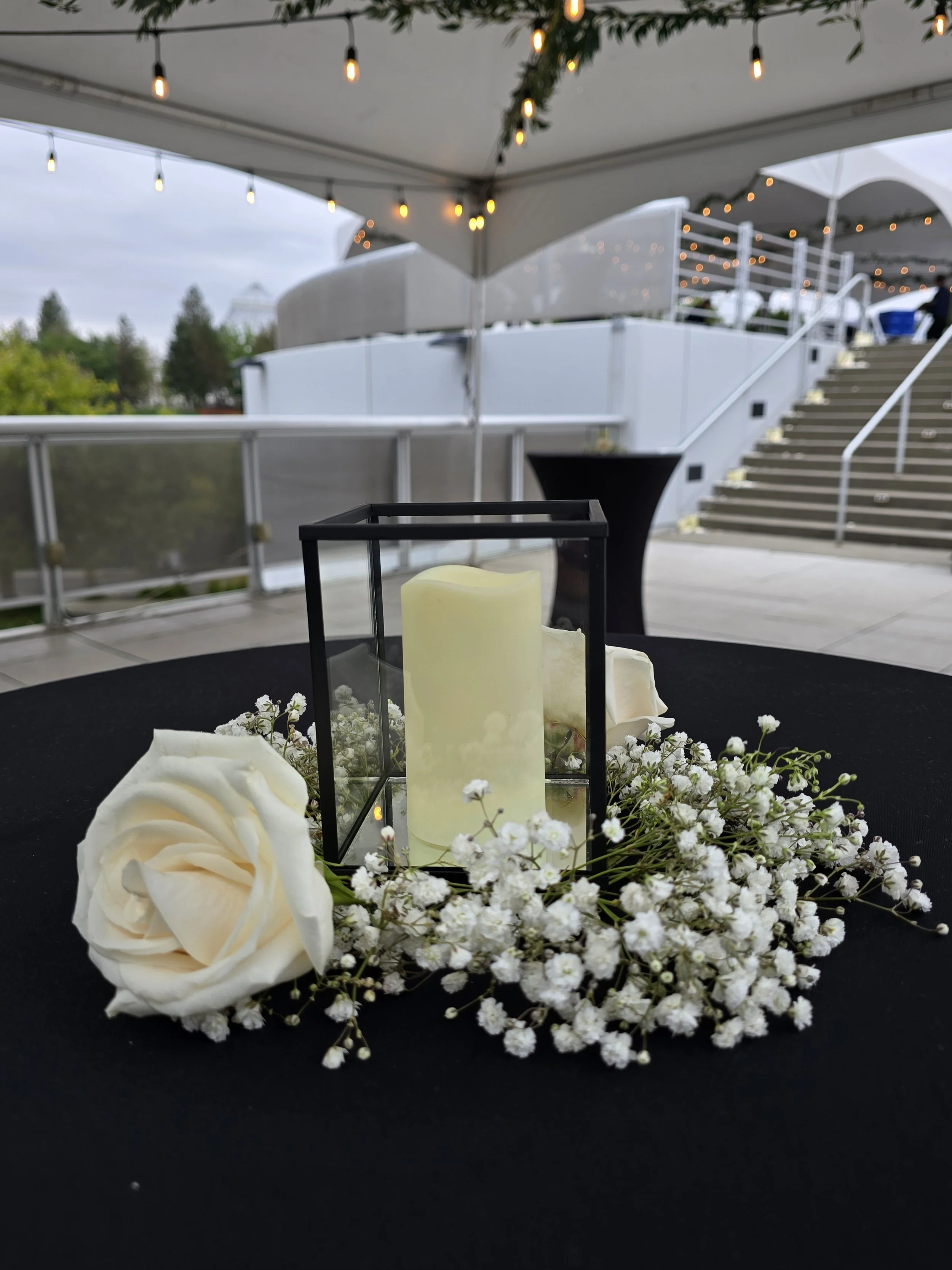 A table decorated with white flowers, including roses and baby's breath, and a candle inside a black metal frame, at an outdoor event under a tent with string lights.