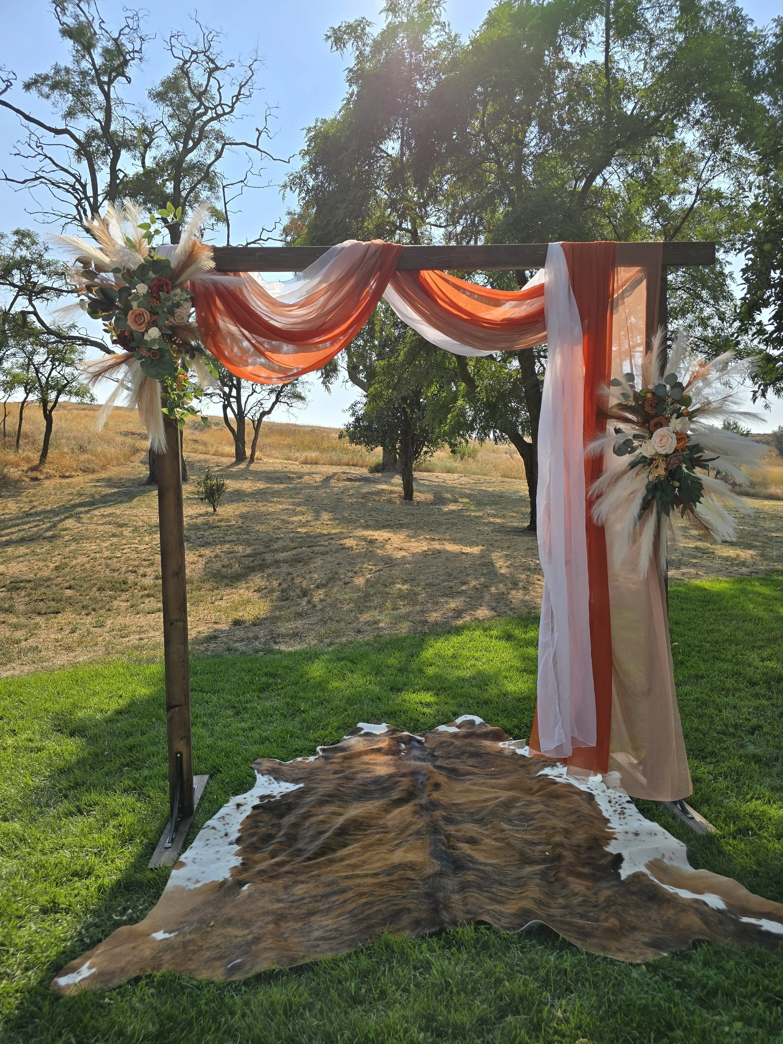 Outdoor wedding arch decorated with orange and white sheer fabric and floral arrangements with roses, eucalyptus, and pampas grass, set on grass with a cowhide rug underneath, trees and open field in the background.