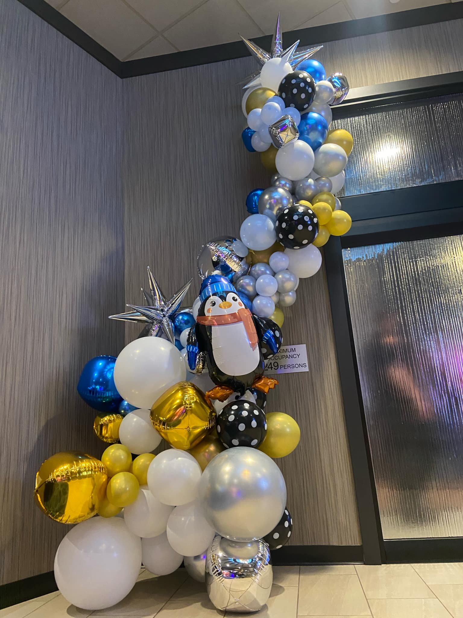 A balloon arrangement featuring gold, silver, white, blue, and black balloons, with some decorated with polka dots and metallic finishes. The arrangement has star-shaped balloons and a large penguin-shaped balloon at the center, with a small sign ind