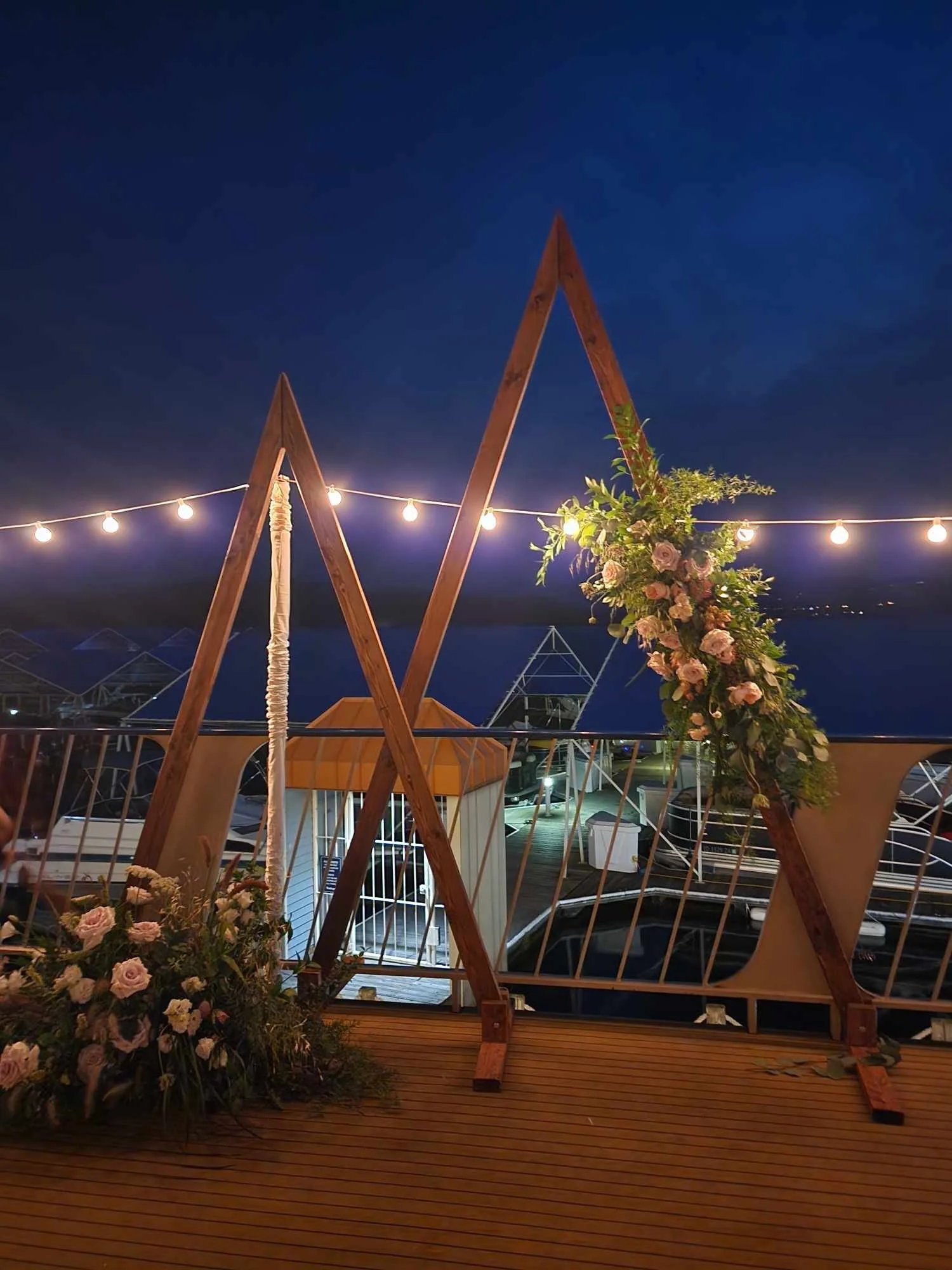 Decorative wooden triangles adorned with floral arrangements and string lights at night over marina with boats.