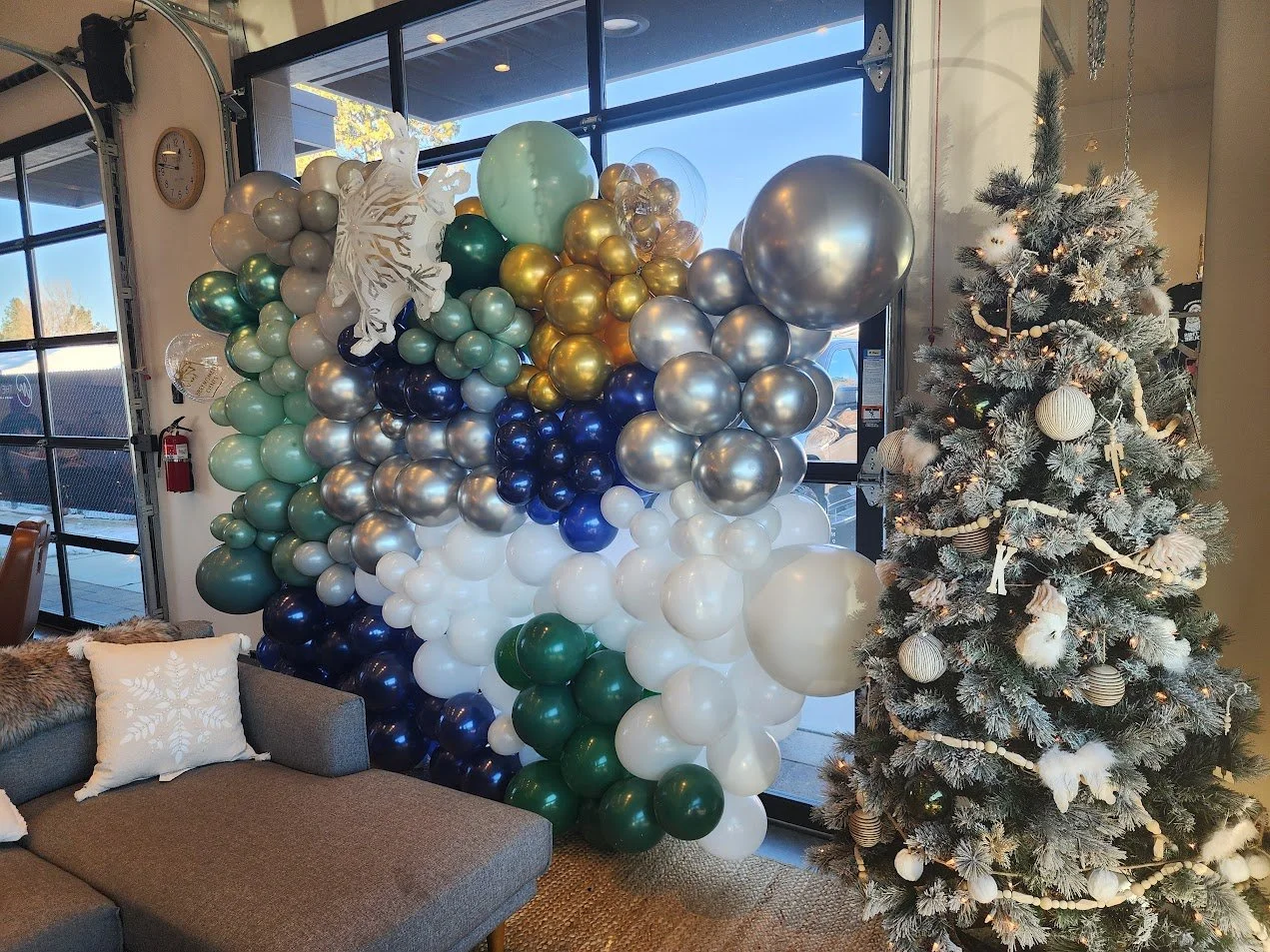 Decorated Christmas tree with white and gold ornaments next to a balloon display with various colored balloons, including white, blue, green, gold, silver, and navy, in a room with large windows.