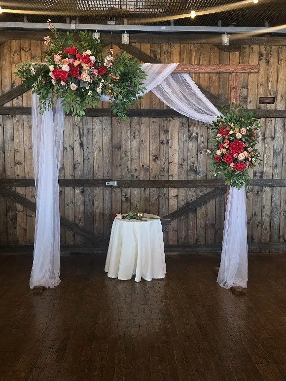 Wedding arch decorated with pink, red, and white flowers, white sheer fabric draping, against a rustic wooden wall, with a small round table covered in white cloth underneath.