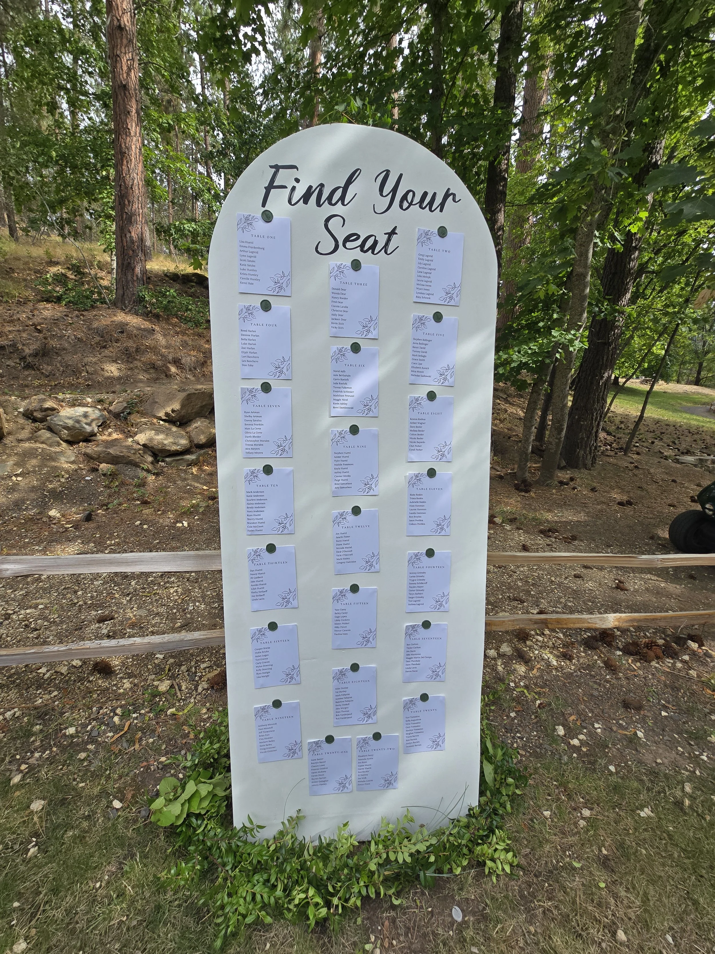 A large white sign with the heading 'Find Your Seat' surrounded by trees and greenery. The sign has multiple sheets of paper attached with seating arrangements for a table event.
