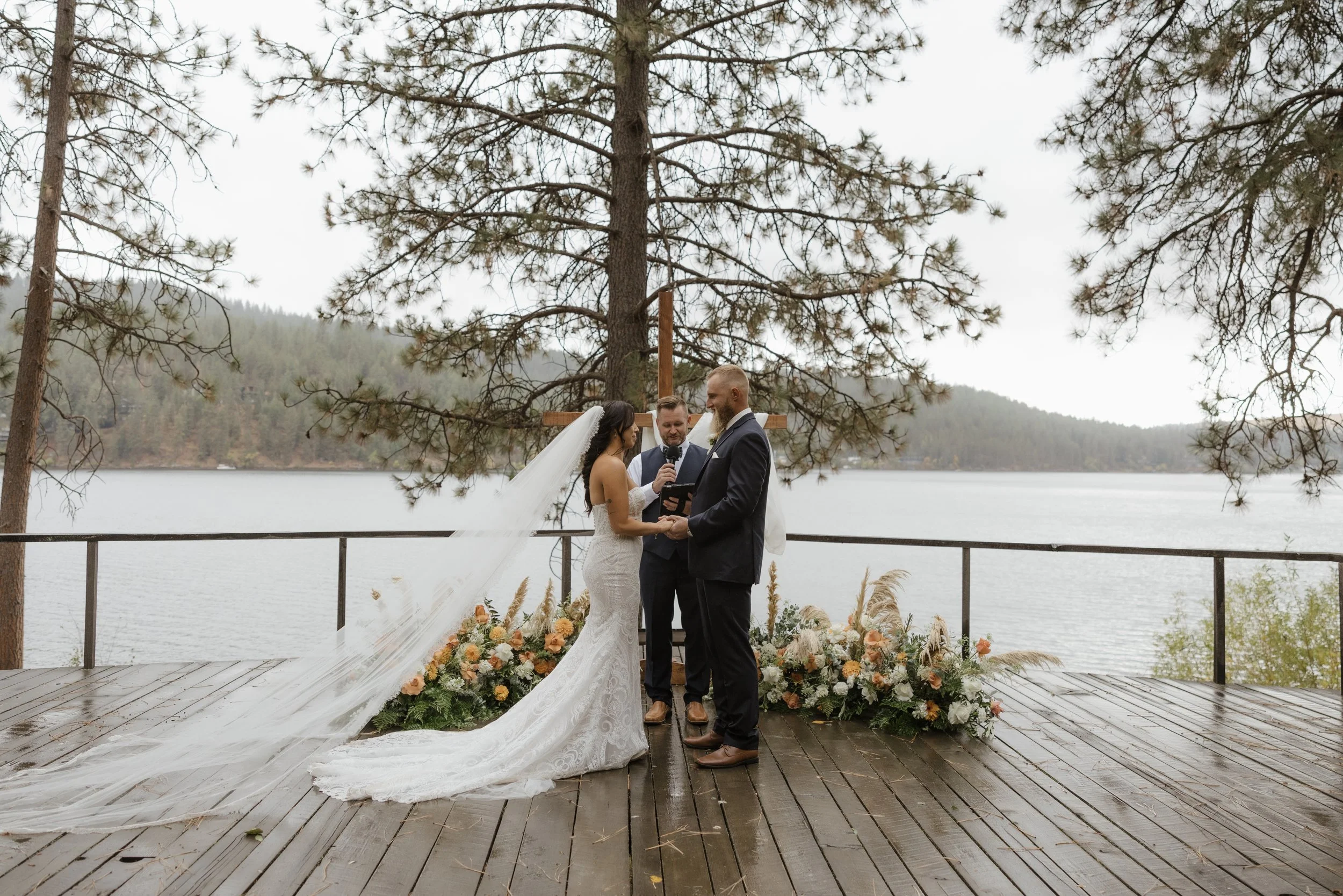 A wedding ceremony taking place outdoors on a wooden platform beside a lake, with a bride and groom holding hands in front of a minister, beneath a large tree and floral arrangements.