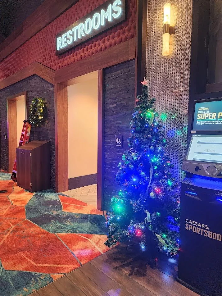 Decorated Christmas tree with colorful lights near a Caesars Sportsbook kiosk in what appears to be an indoor casino or entertainment venue, with a restroom sign and holiday wreath visible in the background.
