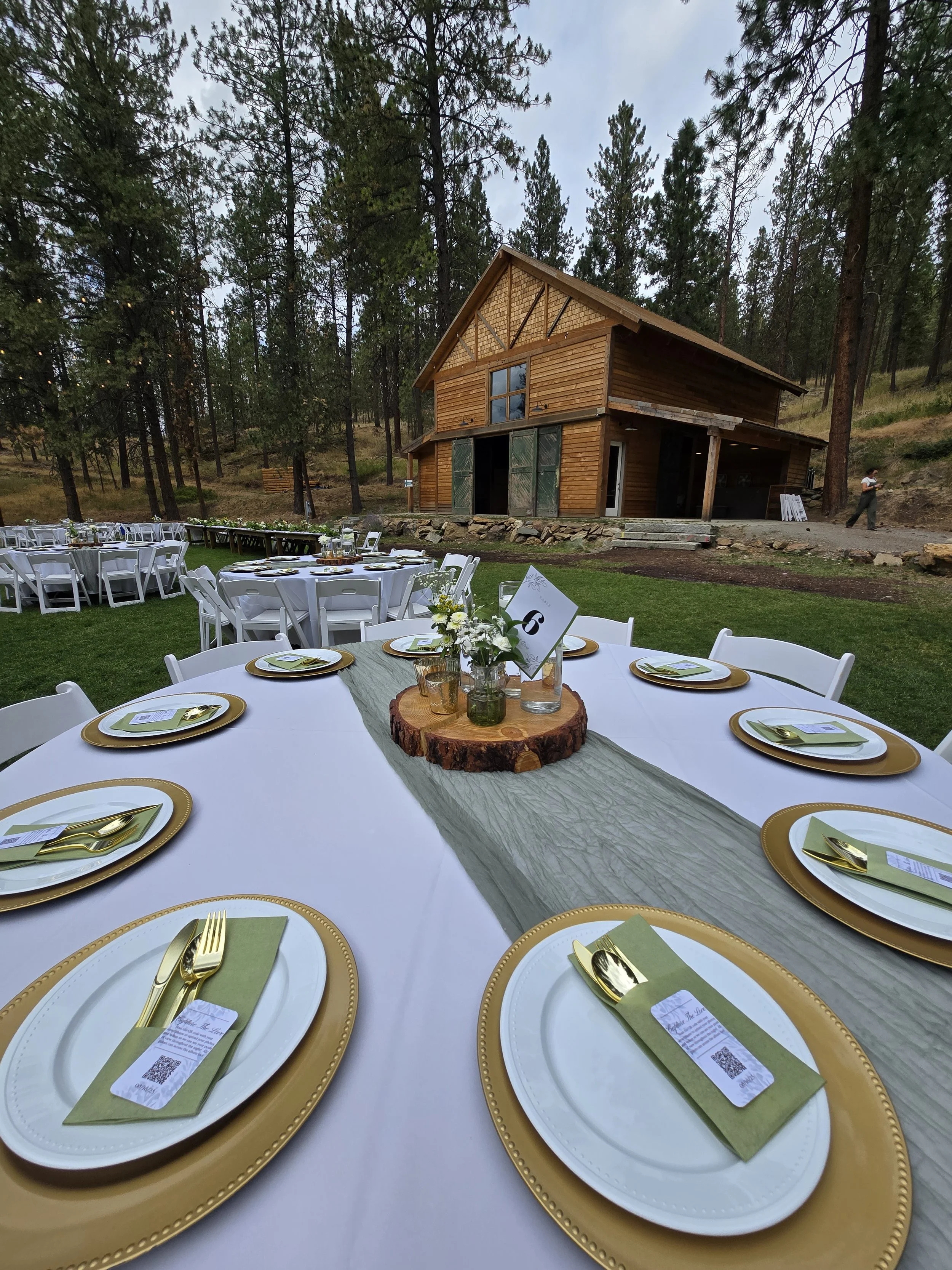 Outdoor event setup with round tables covered in white tablecloths, each with gold and white plates, gold utensils on green napkins, and small floral centerpieces on logs, in front of a rustic wooden house surrounded by trees.