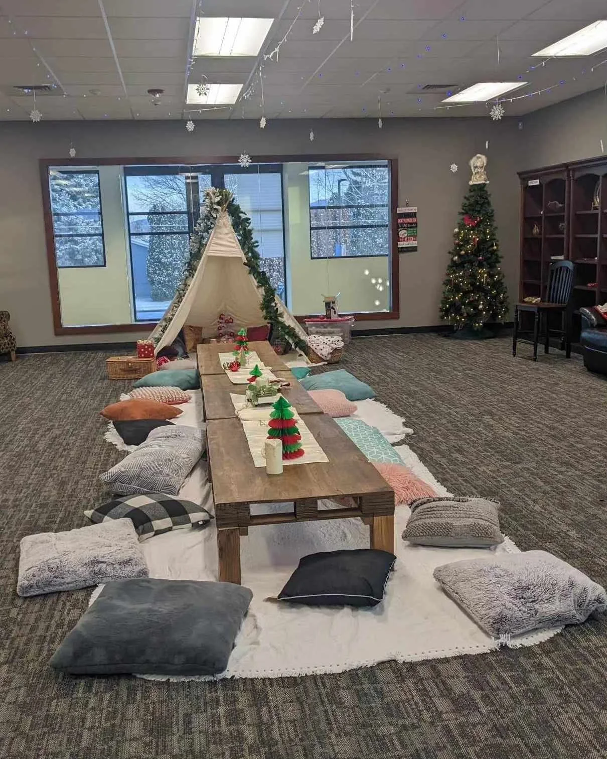 Indoor holiday scene with a teepee tent decorated with greenery, a long wooden table with small Christmas trees and candles, surrounded by cushions and rugs, Christmas tree with ornaments and a star on top, snowflake decorations hanging from the ceil