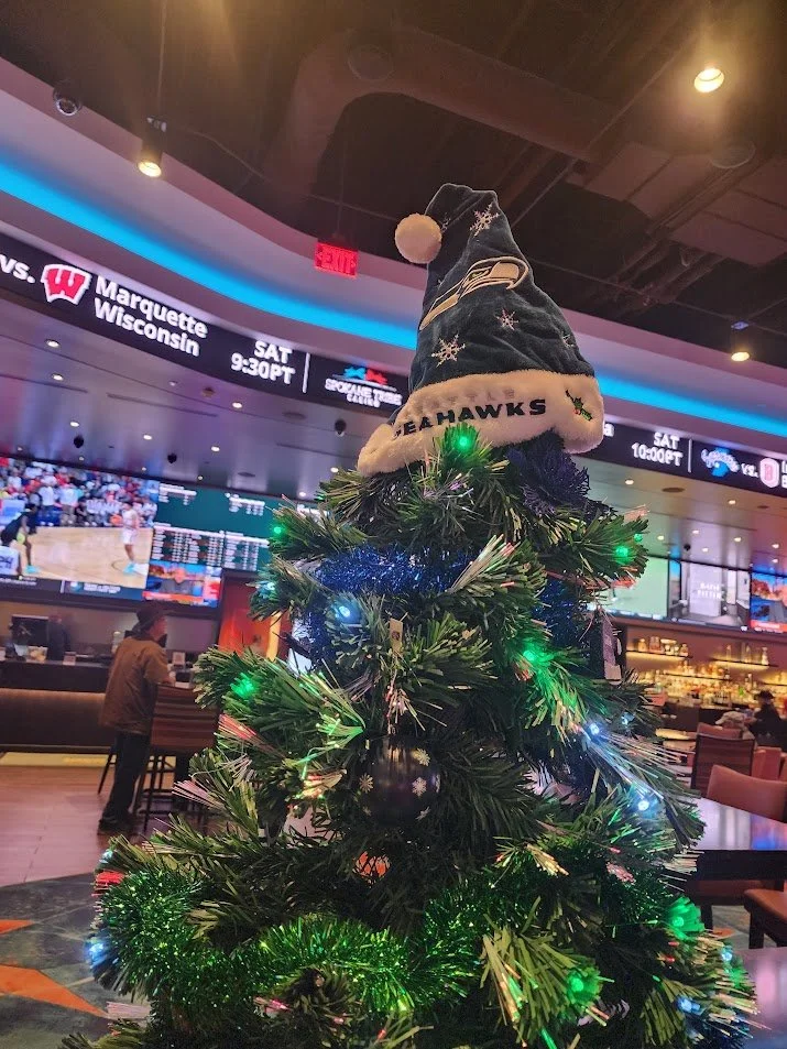 Small decorated Christmas tree with a Seattle Seahawks Santa hat on top, set inside a sports bar or restaurant with TV screens and other people in the background.