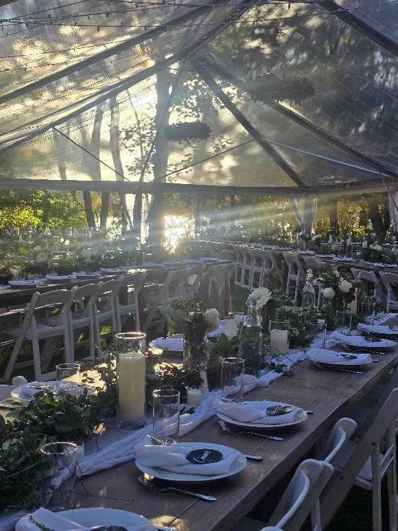 Long dining table set with white plates, silverware, candles, and greenery in a tented outdoor setting with sunlight filtering through trees.