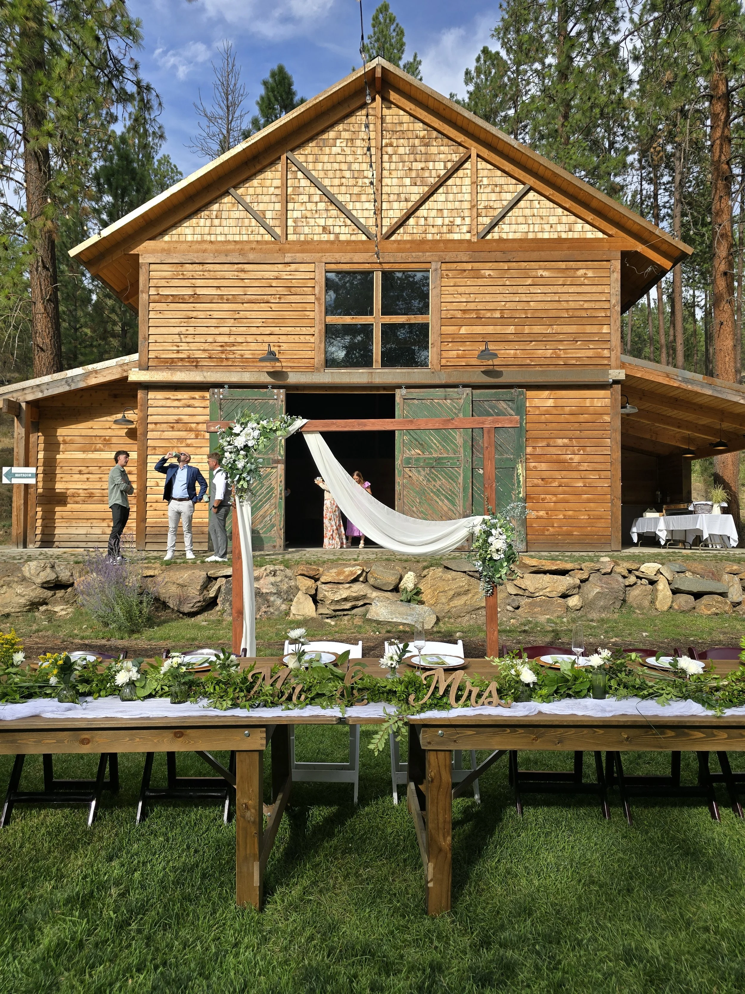 A rustic wooden barn is decorated for a wedding with a white draped fabric and floral arrangements. There is a table in the foreground with floral decorations and place settings. A few guests are standing near the barn entrance, which has open green 