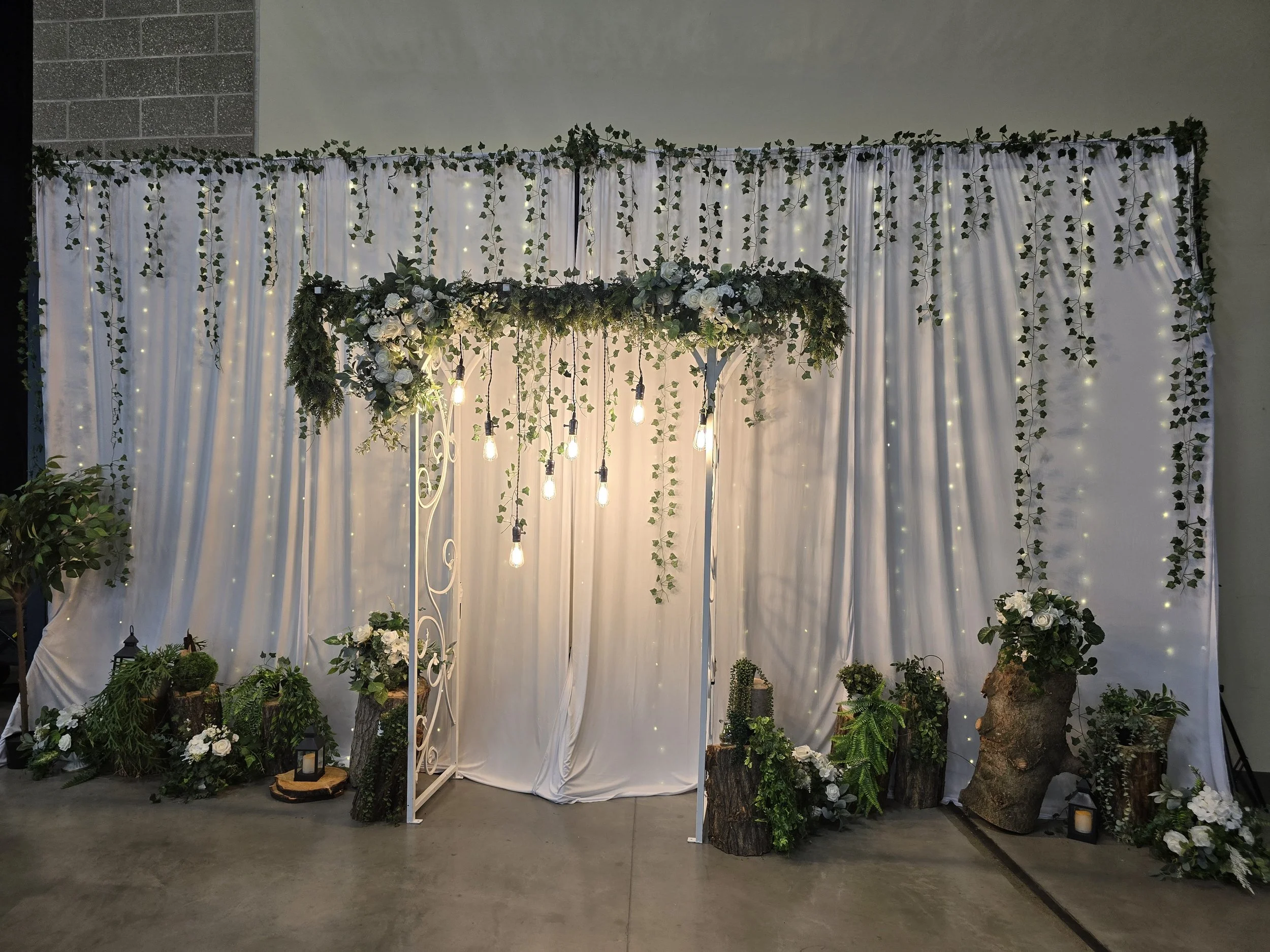 Decorative wedding or event backdrop with white fabric curtains, hanging string lights, and greenery, including potted plants, logs, and lanterns at the base.