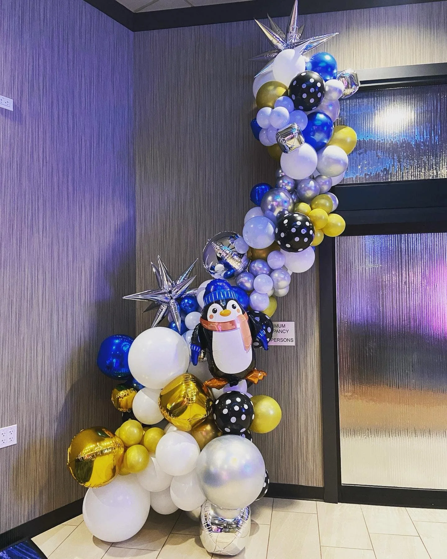 Balloon arrangement with white, black, gold, blue, and iridescent balloons, including star-shaped balloons and a penguin-shaped balloon, set against a textured wall near a glass door.